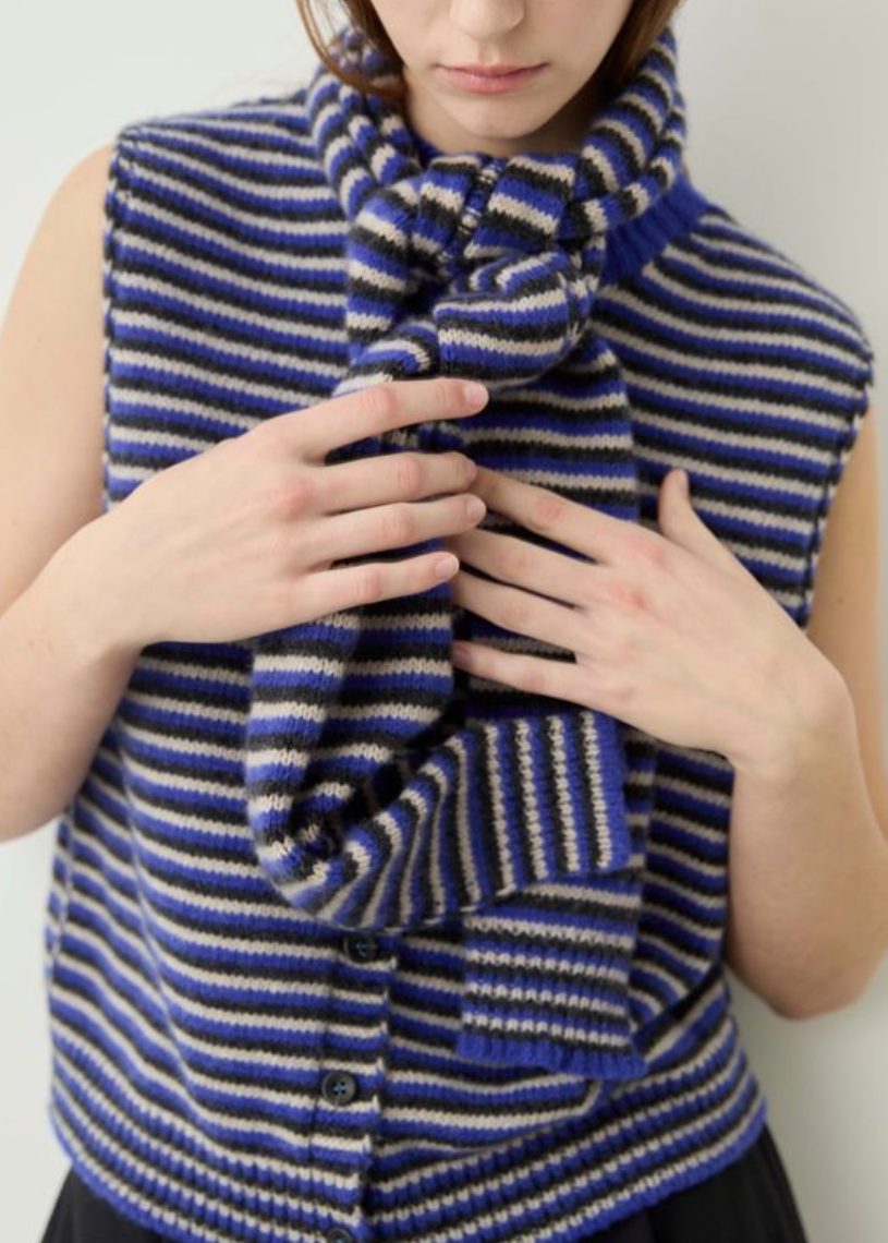 A close-up of a person wearing a sleeveless knit cardigan vest with blue, navy, and beige horizontal stripes, paired with a matching striped knit scarf.