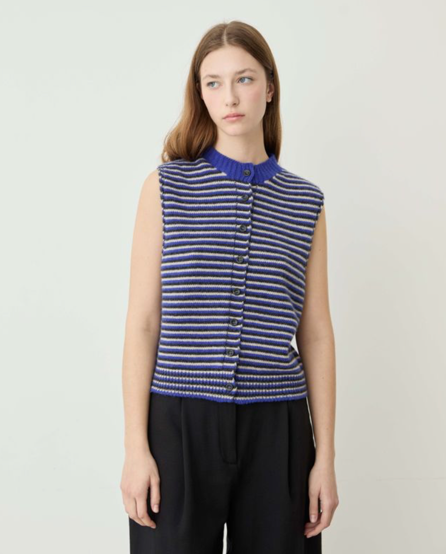 A woman modeling a sleeveless ribbed knit vest with horizontal blue, navy, and white stripes, featuring a button-up front and a mock neck, paired with black trousers against a light gray studio background.