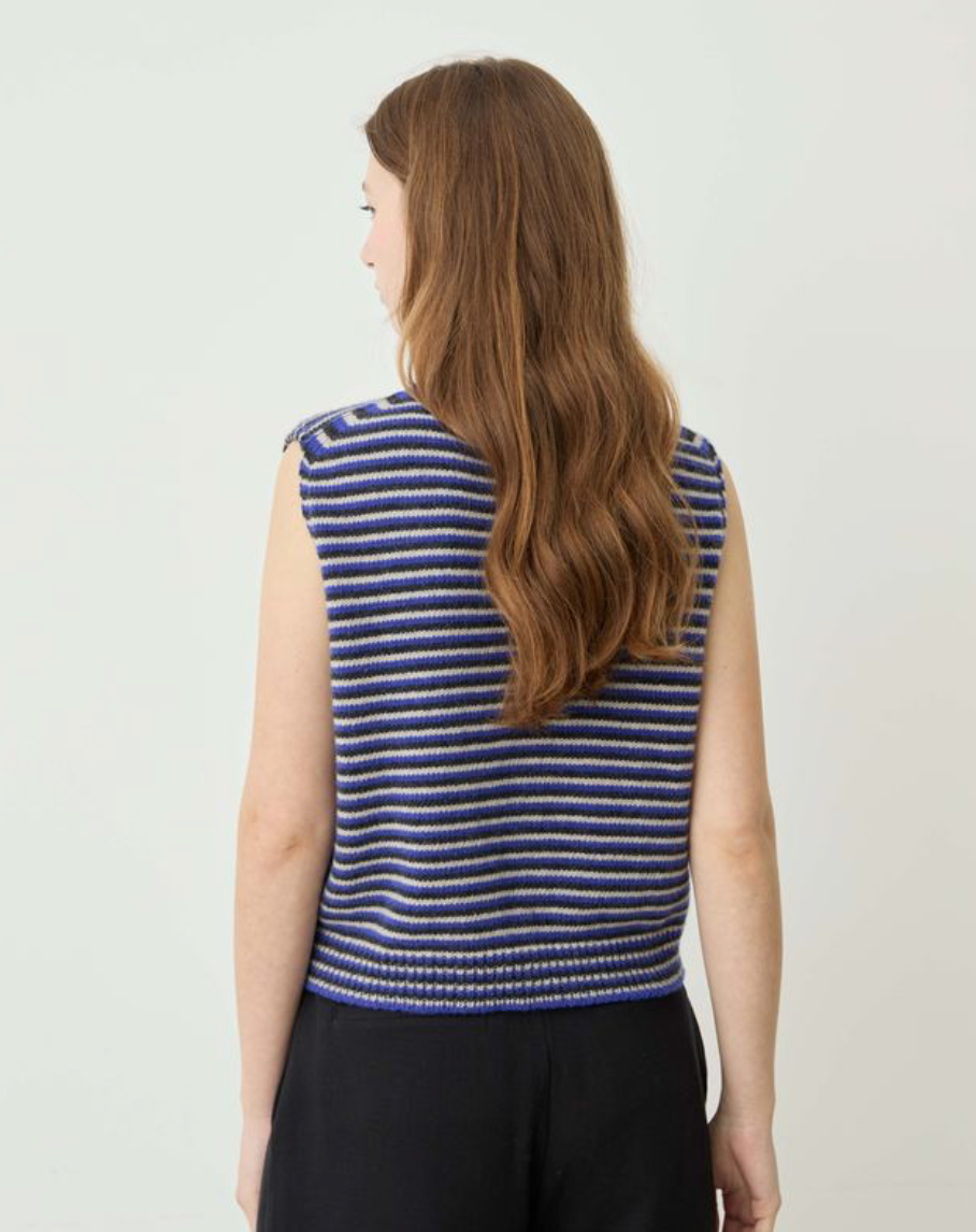 A woman with long wavy brown hair is shown from behind wearing a sleeveless knit top with horizontal blue, navy, and white stripes, paired with black pants, against a plain light gray background.