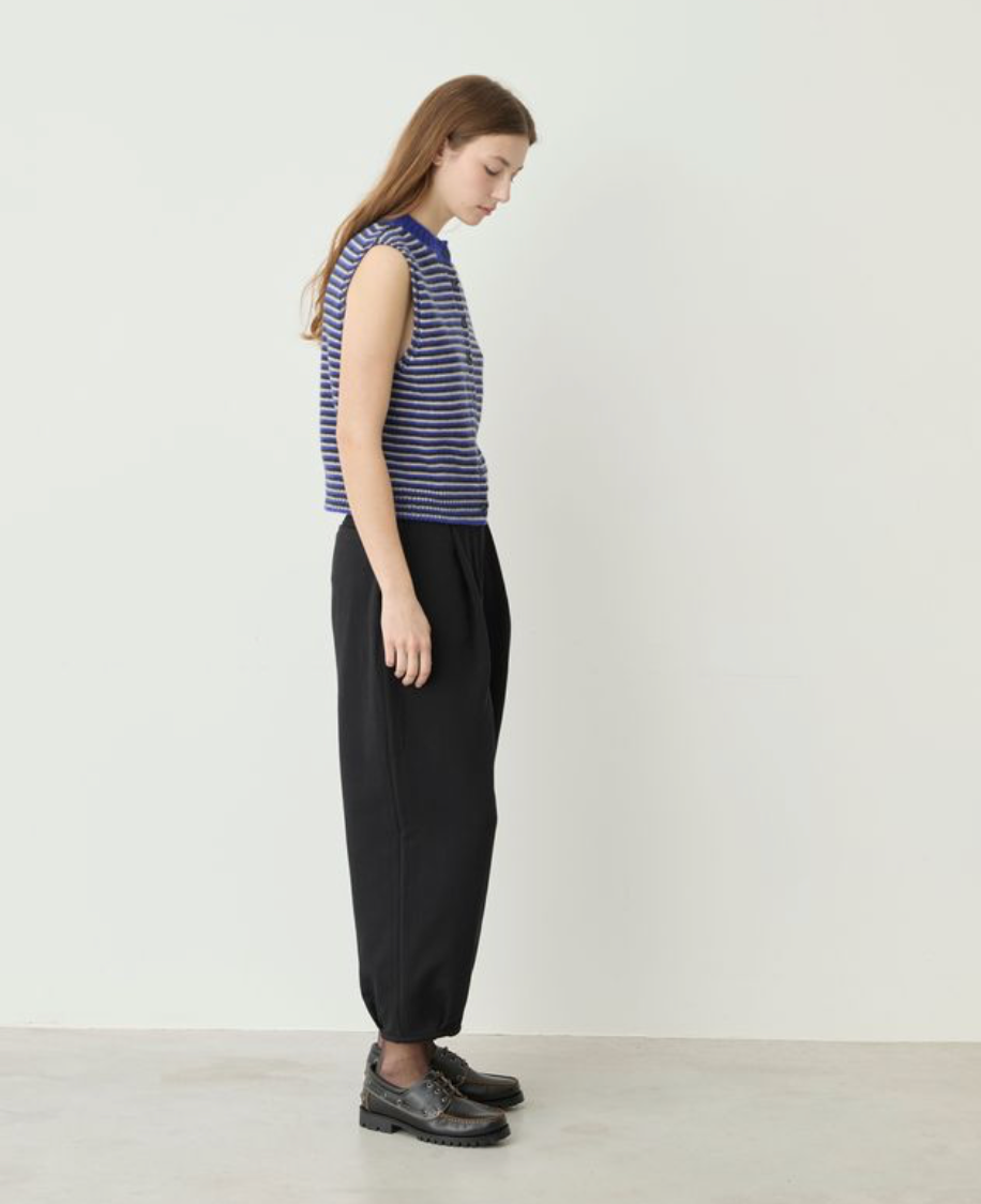 A woman stands in profile wearing a sleeveless top with thin blue and white horizontal stripes and loose black pants, against a plain off-white studio wall.