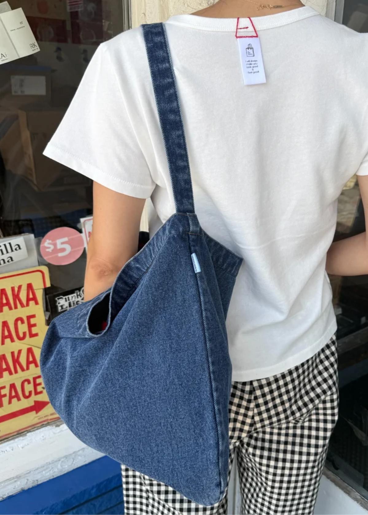 A person wearing a white t-shirt and black-and-white gingham pants carries a large, slouchy blue denim shoulder bag with a wide strap in front of a shop window.