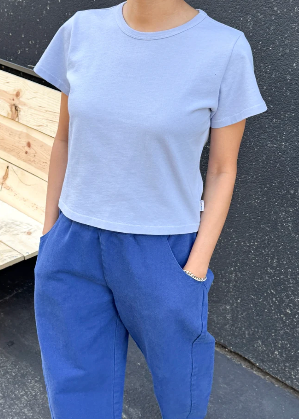A person wearing a light periwinkle blue short-sleeve crewneck t-shirt and blue pants, standing outdoors against a dark textured wall.