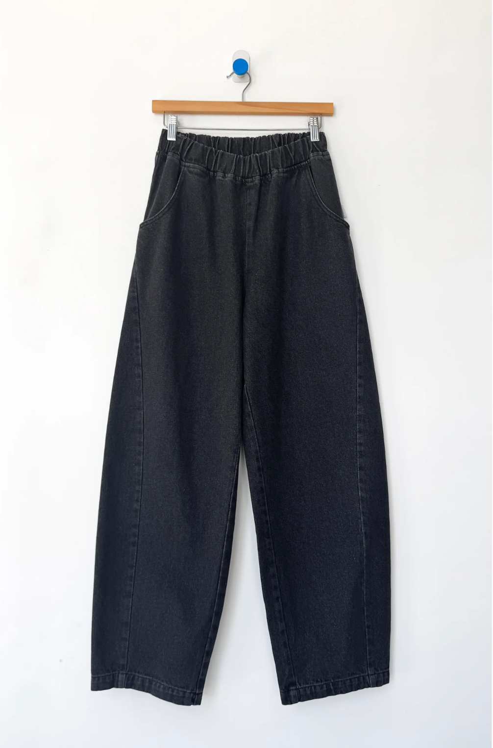 Black wide-leg cropped jeans with an elastic waistband, hanging from a wooden hanger against a plain white wall.