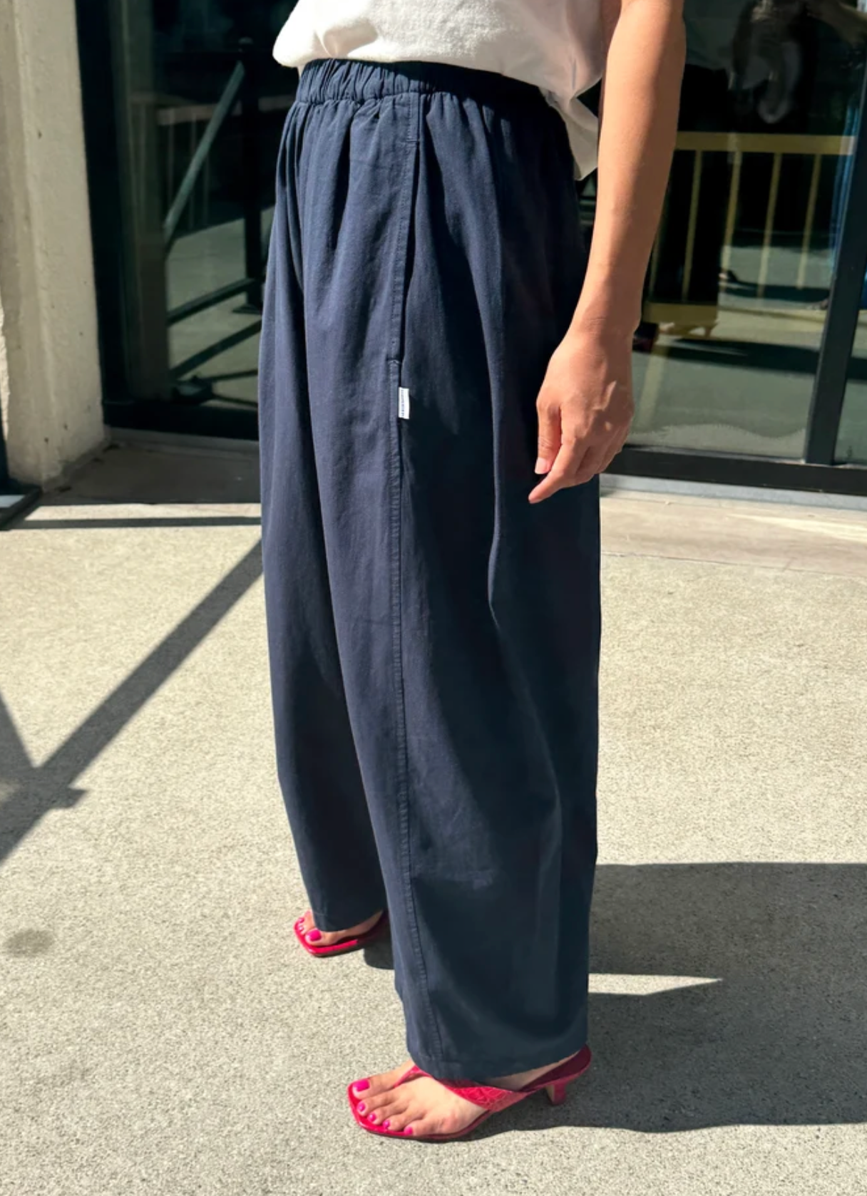 A person wearing loose, wide-leg navy pants with an elastic waistband, paired with a white top and pink heeled sandals, standing outdoors on a concrete surface.