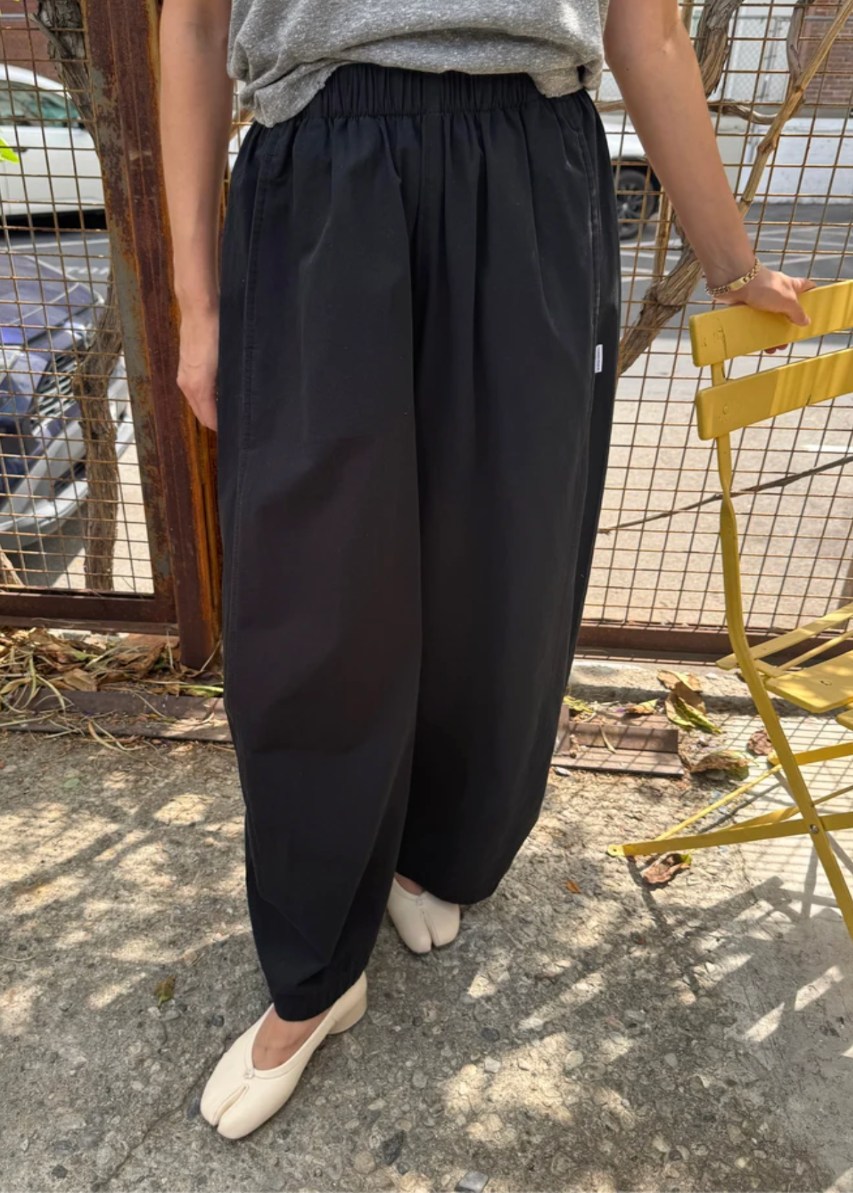 A person wearing loose black wide-leg pants with an elastic waist, paired with a gray top and cream-colored shoes, standing outdoors on a concrete surface next to a yellow chair.