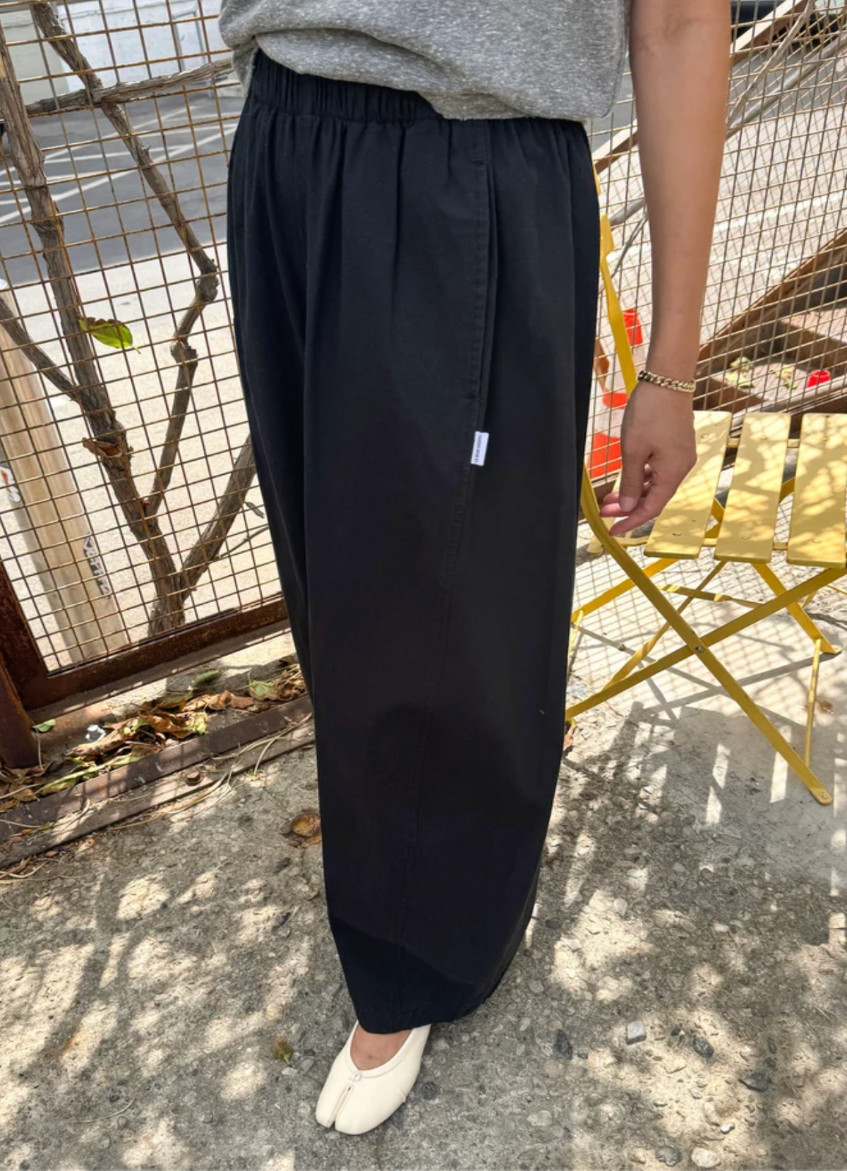 A person wearing loose black wide-leg pants with an elastic waistband, paired with a gray top and white tabi-style shoes, standing outdoors on a concrete surface near a yellow folding chair.
