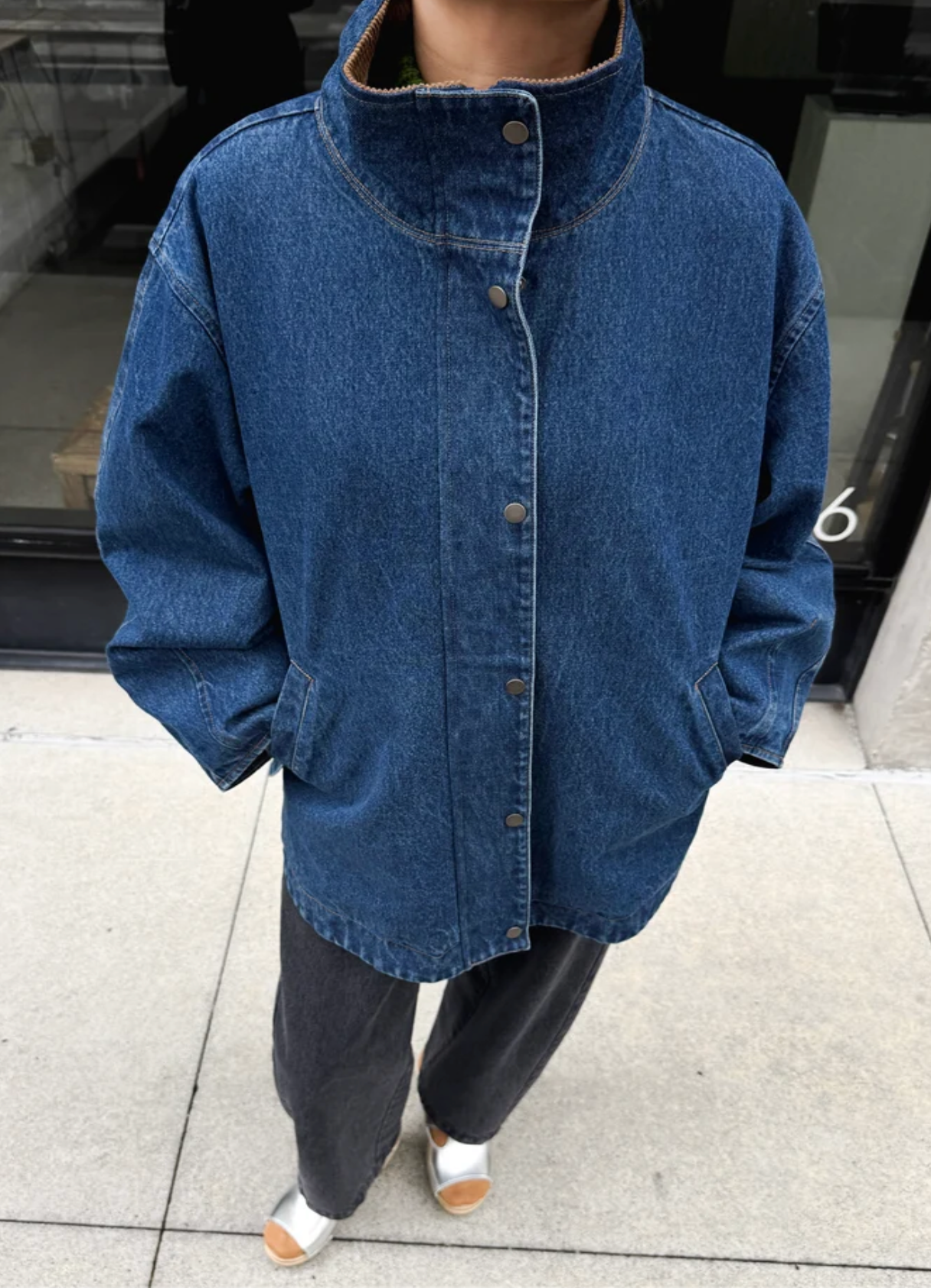 A person wearing an oversized blue denim jacket with a high stand collar and front buttons, paired with dark pants and white shoes, standing outdoors on a tiled walkway.