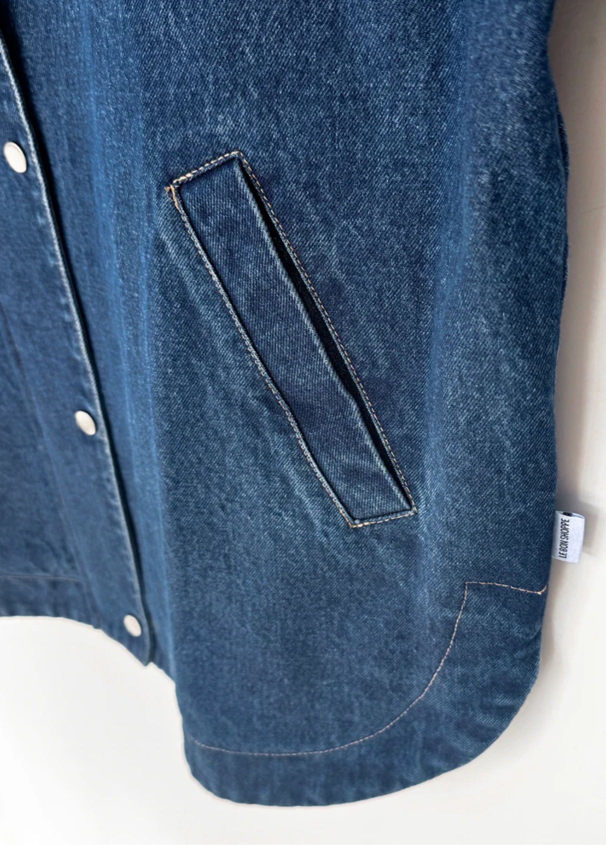 Close-up of a blue denim jacket showing a diagonal welt pocket, white buttons, and a small white brand tag 