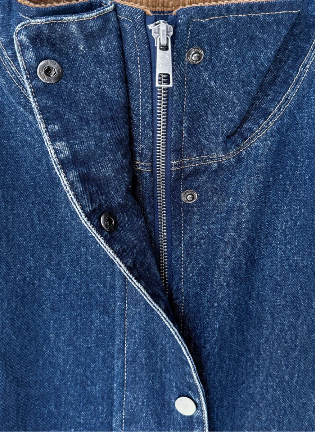 Close-up of a blue denim garment showing the front closure with a central silver zipper, metal snap buttons, and contrast stitching.