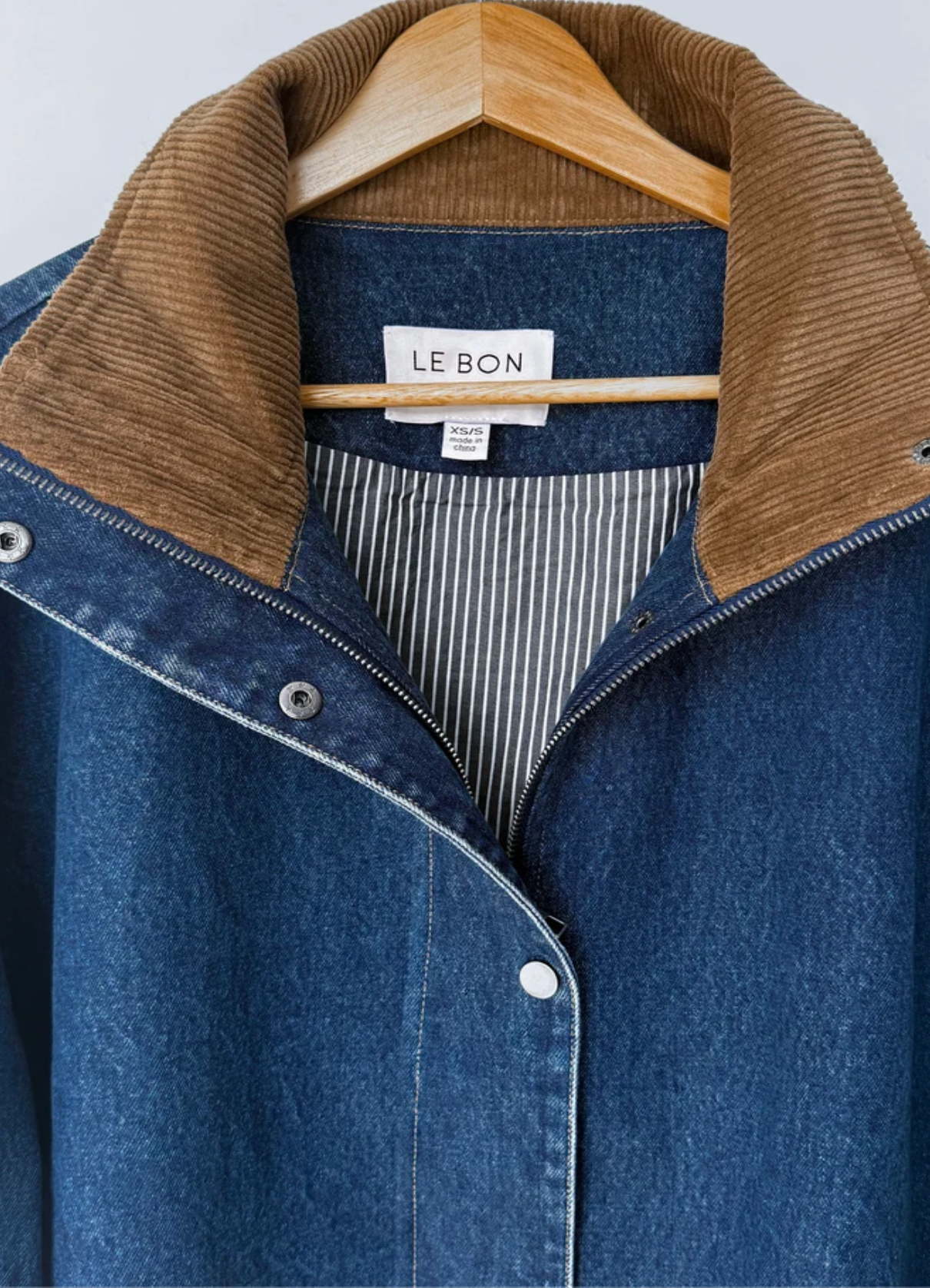 A close-up of a blue denim jacket with a brown corduroy collar, hanging on a wooden hanger. The brand label 'LE BON' is visible inside the collar.