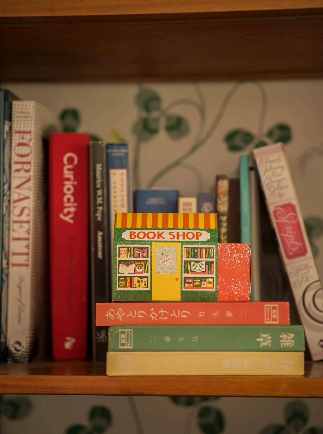A miniature toy bookshop set styled on a wooden bookshelf among real books. The toy set includes a small storefront with a red and yellow striped awning, colorful windows, and a green roof, alongside three matching books stacked in front.