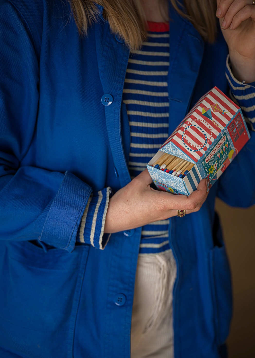 illlustrated seaside match box with stripes ice creams and spades in the window a red door and more beachy drawings being held by a model wearing a striped blue and white shirt and a cobalt blue core jacket