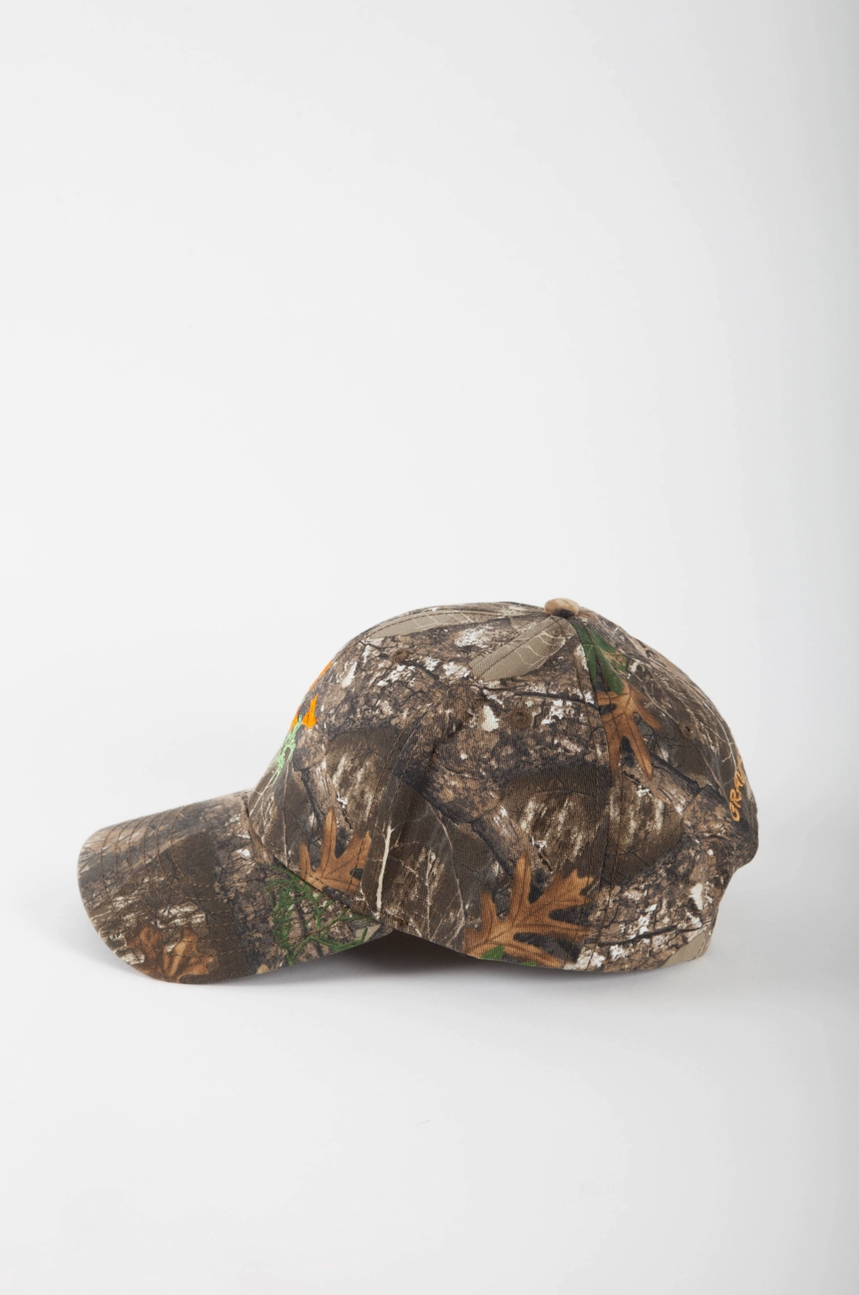 A baseball cap in a realistic woodland camouflage pattern featuring brown leaves, green branches, and gray tree bark, shown from a side profile against a light gray background.
