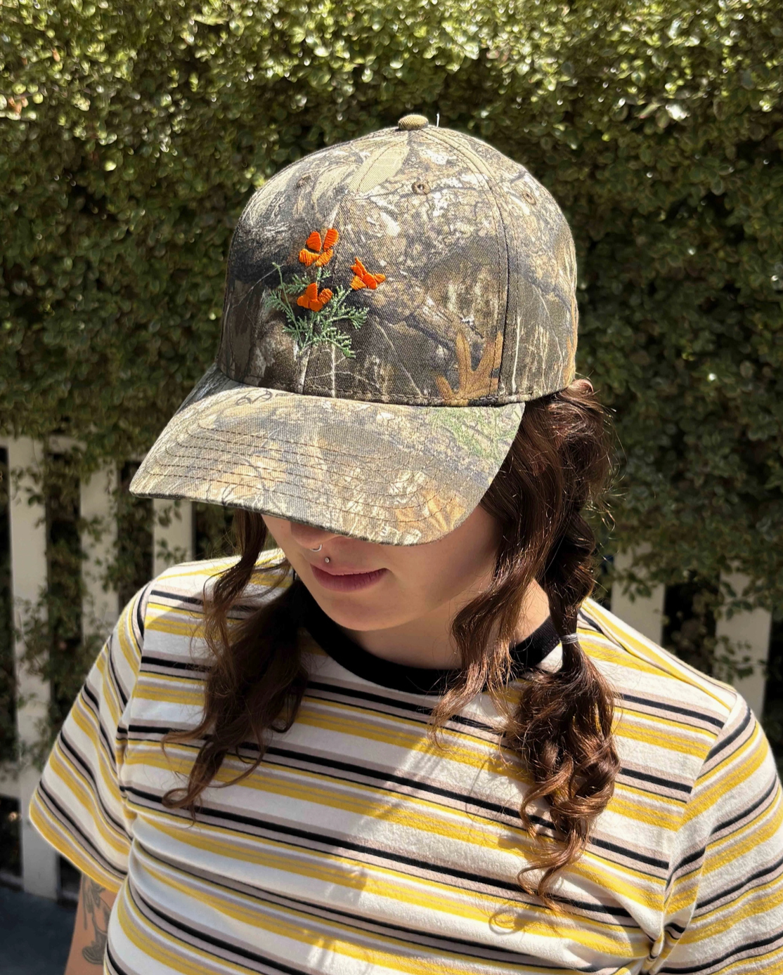 A person outdoors wearing a camouflage baseball cap with embroidered orange flowers and green leaves on the front, paired with a yellow, white, and black striped shirt.