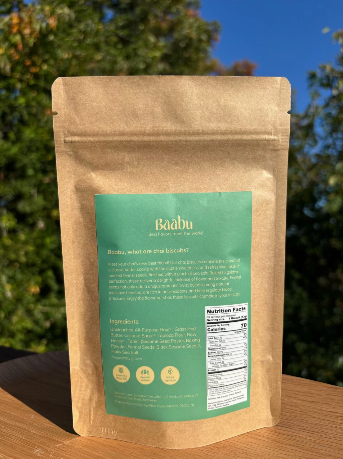 A kraft paper stand-up pouch of Baabu choi biscuits with a large green label, photographed outdoors on a wooden surface with trees and blue sky in the background.