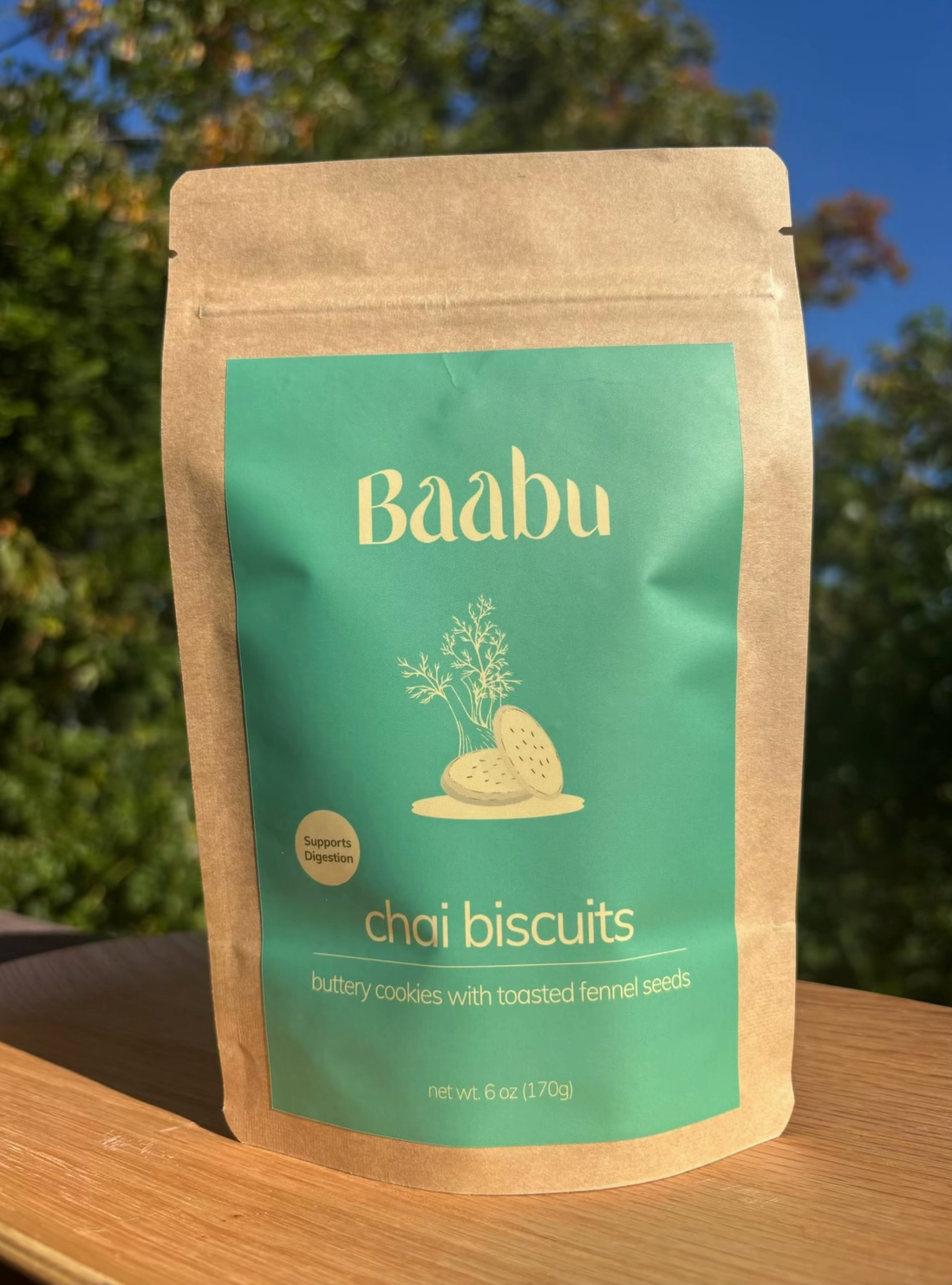 A stand-up kraft paper pouch of Baabu Chai Biscuits placed on a wooden surface outdoors, with blurred green trees and blue sky in the background.