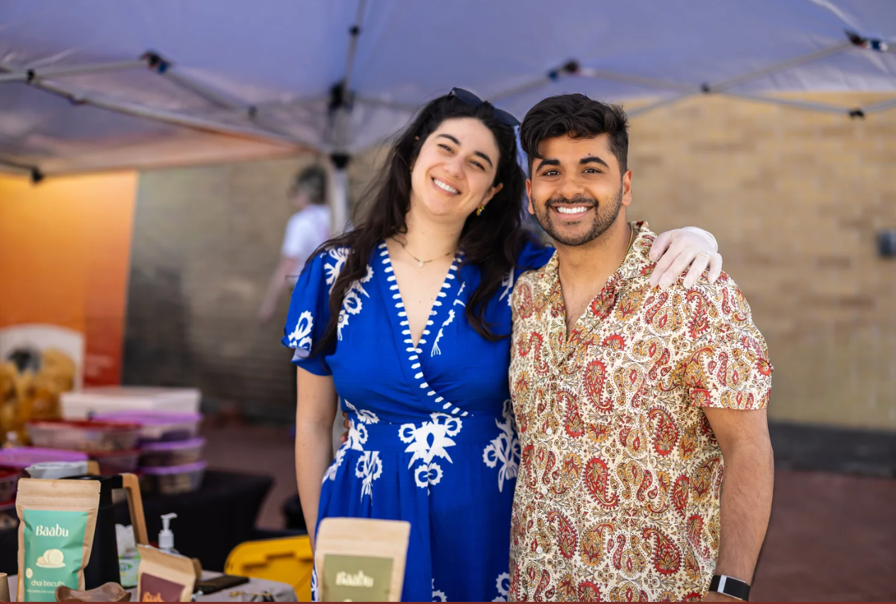 A smiling couple standing together at an outdoor market booth under a canopy tent. The woman wears a bright blue dress with white embroidery, and the man wears a beige short-sleeve button-up shirt with a red paisley pattern.