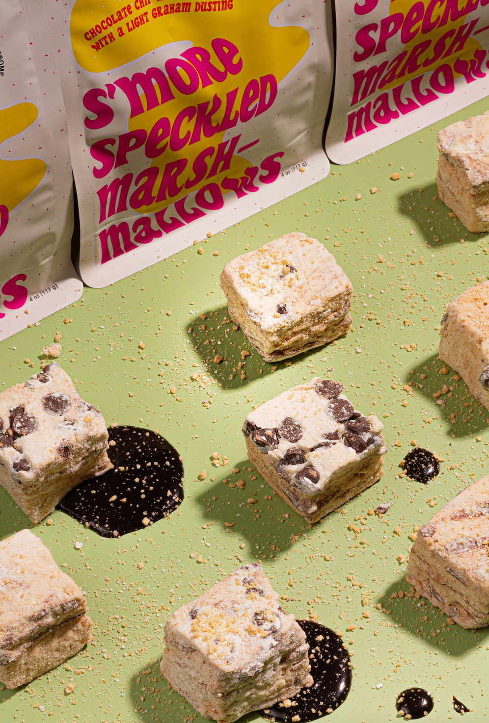 Several square chocolate chip marshmallow treats are arranged on a light green surface with dark chocolate drizzle, alongside branded stand-up pouches reading 'S'more Speckled Marshmallows.' 