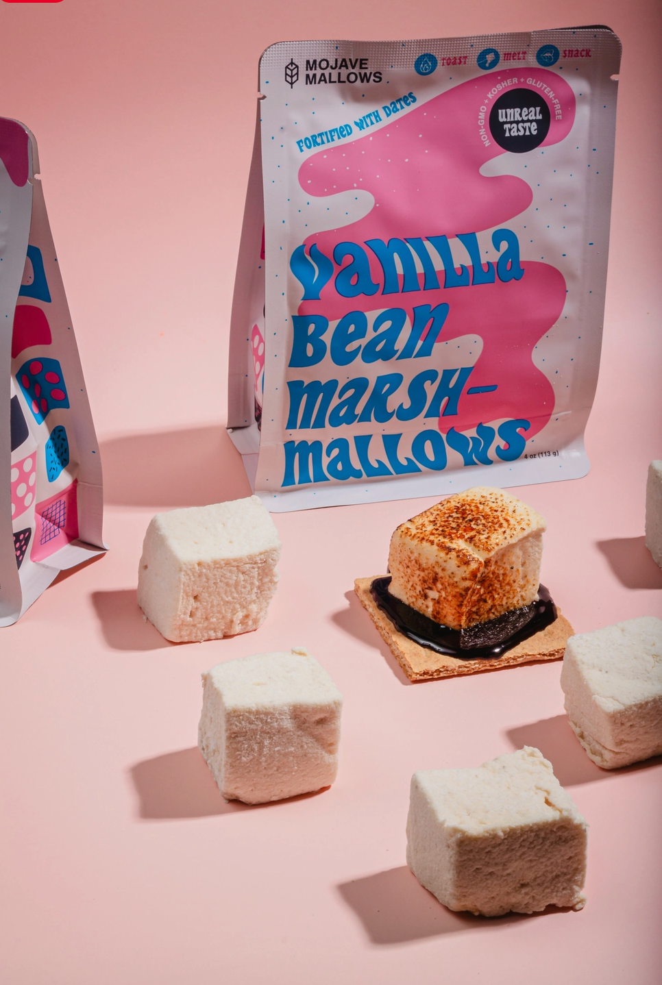 Mojave Marshmallows vanilla bean marshmallows snack pouch shown with several raw marshmallows and one toasted marshmallow on a graham cracker, arranged on a pink background.