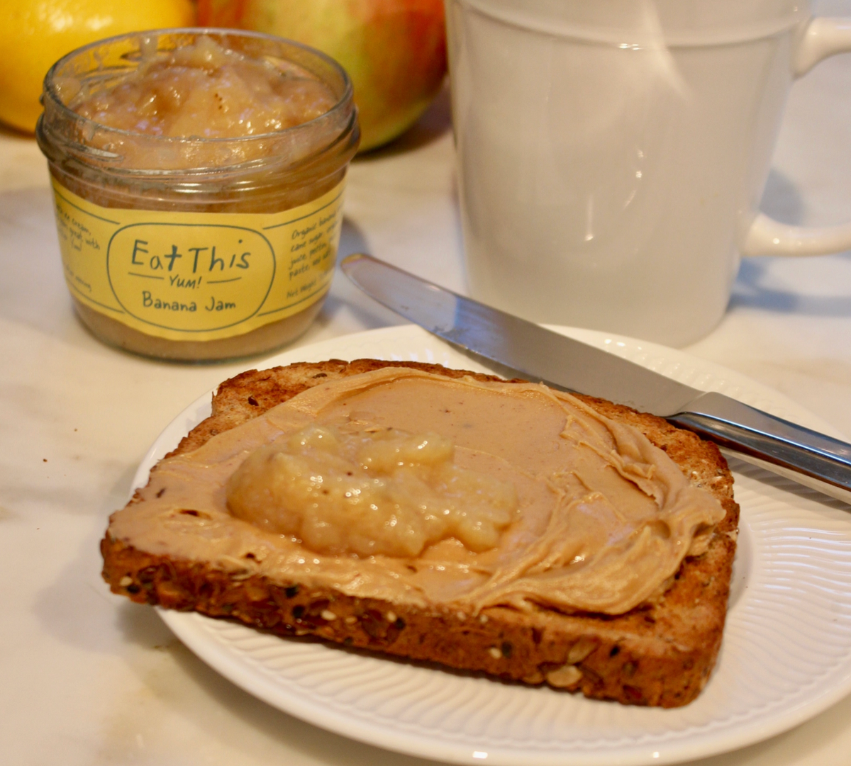 A slice of toast with banana jam spread on it sits on a white plate, next to a jar of Eat This Your Banana Jam and a white mug on a light-colored countertop.