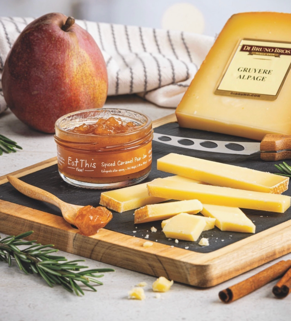 A styled food scene showing a small glass jar of spread on a wooden board with cheese wedges, a pear, cinnamon sticks, and a cloth in the background. The jar label reads Eat This Co and the spread appears amber-orange.