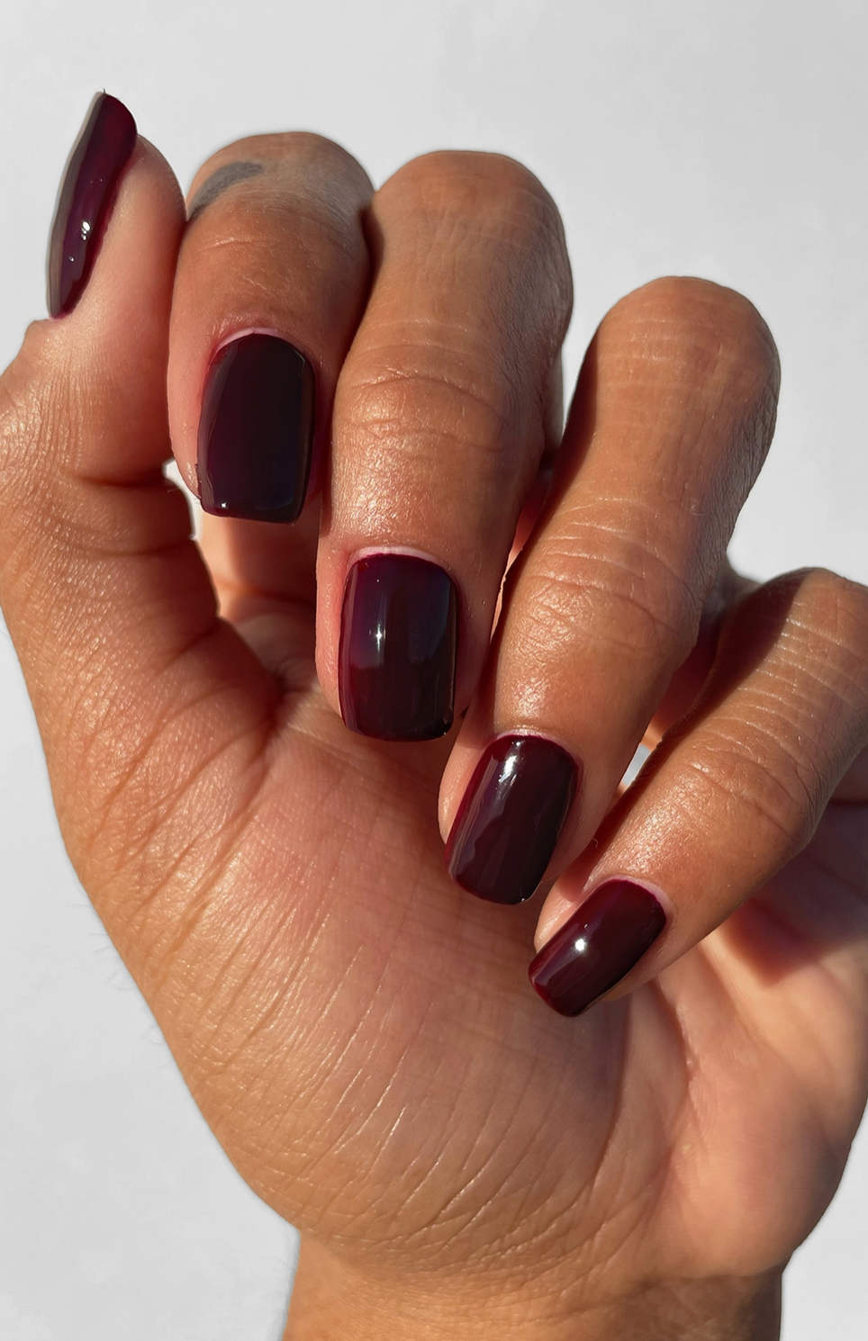 A close-up of a hand with neatly manicured almond-shaped nails painted in a deep, glossy burgundy shade against a plain light background.