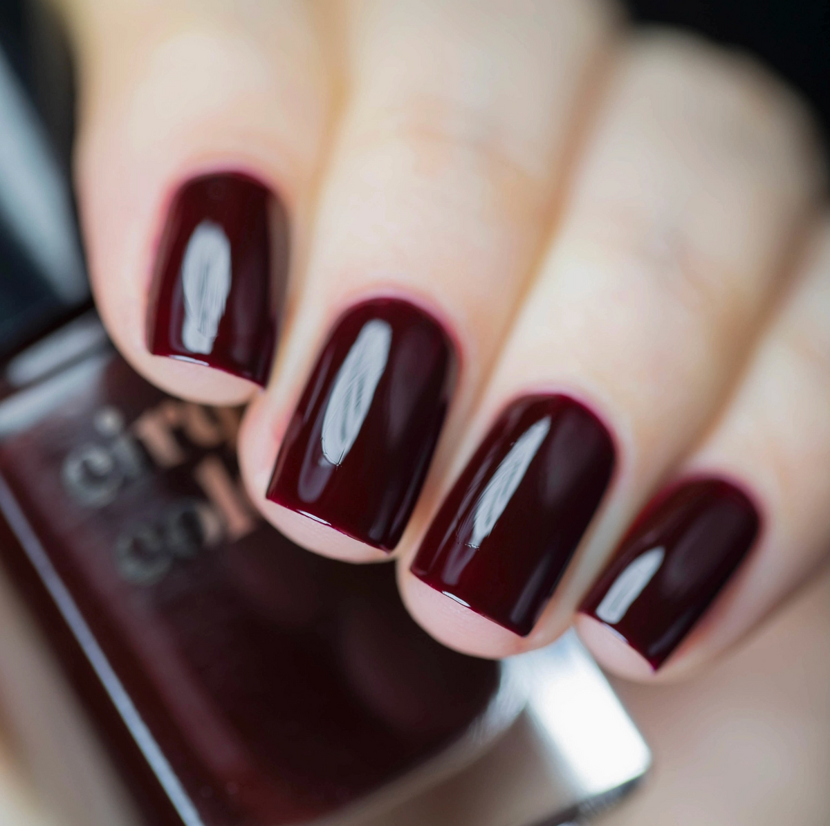 A close-up of a hand with fingernails painted in a glossy deep burgundy nail polish, with a matching bottle of nail polish partially visible in the foreground.