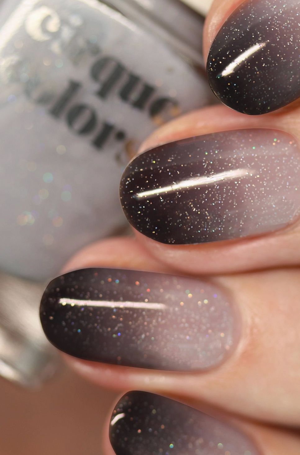 Close-up of fingernails painted with a dark glitter nail polish that fades into a sheer pale lavender-gray at the tips. A bottle of Cirque Color nail polish is blurred in the background.