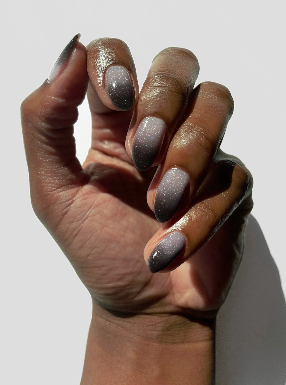 A close-up of a hand with long almond-shaped nails painted in a metallic silver-to-gray ombre glitter manicure, set against a plain light gray background.