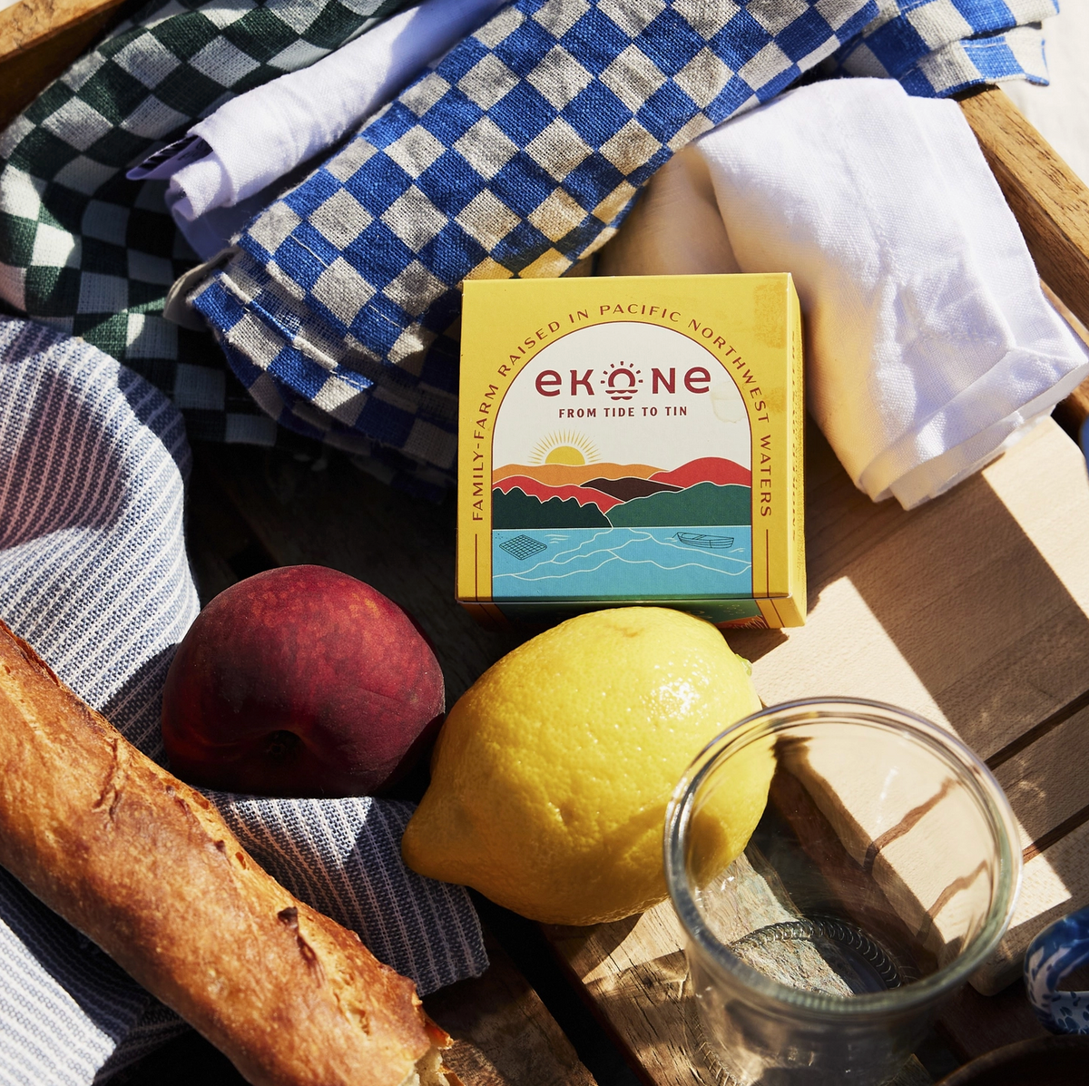 A yellow Ekone box sits on a wooden picnic table surrounded by a blue-and-white checkered blanket, white cloth, striped towel, bread, a peach, and a lemon, with a glass jar in the foreground.