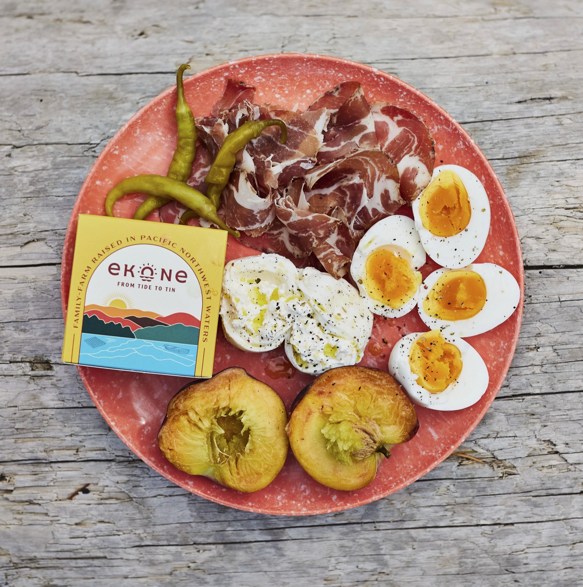 A rustic charcuterie board on a weathered gray wooden table, featuring sliced cured ham, hard-boiled eggs, green beans, halved avocado, and a yellow Eköne box.