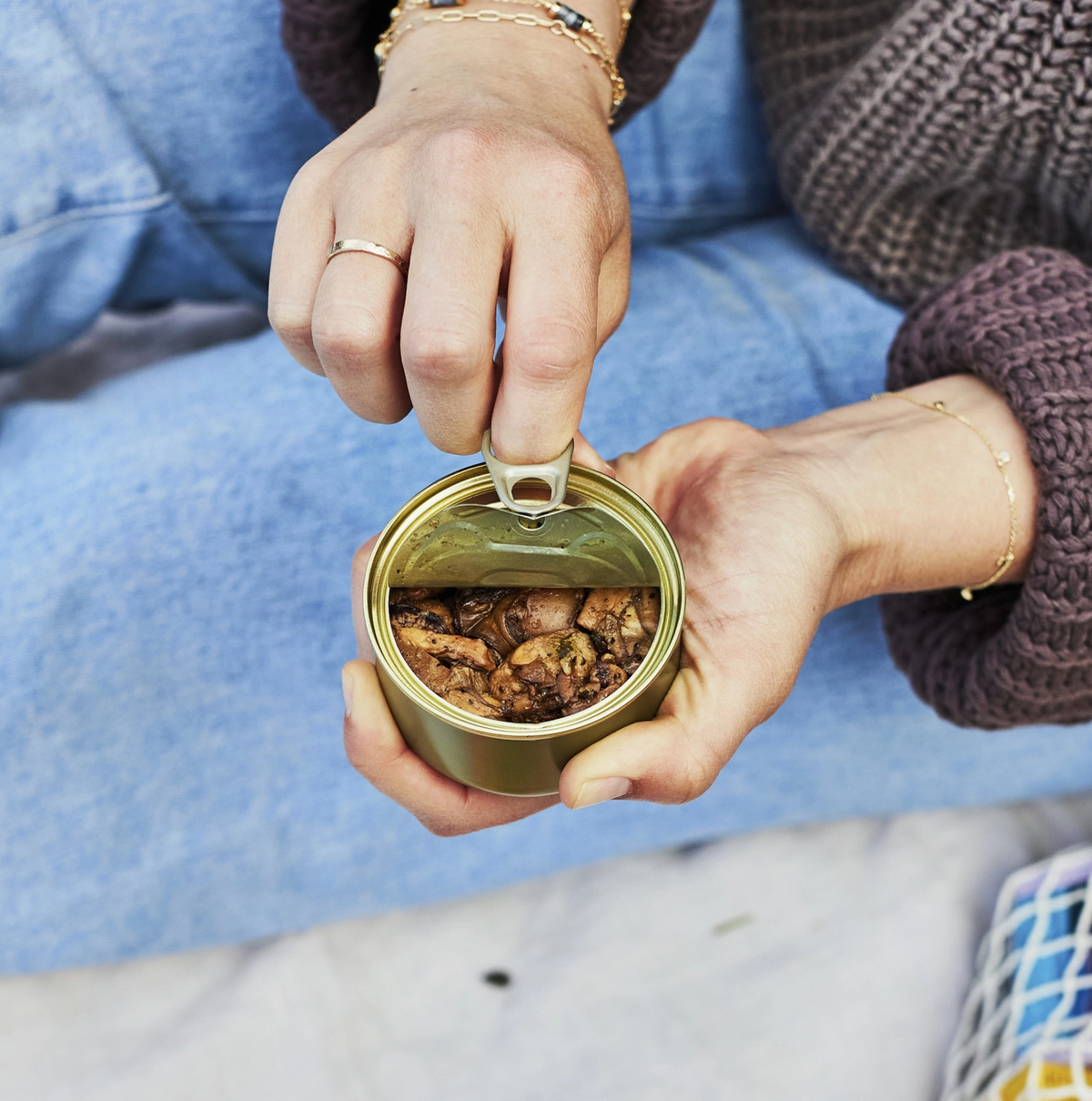 A close-up lifestyle shot of a person's hands opening a small metal tin of nuts with a pull-tab lid. The person is wearing a blue denim jacket and a dark brown knit sweater, with gold jewelry visible on their fingers and wrist.