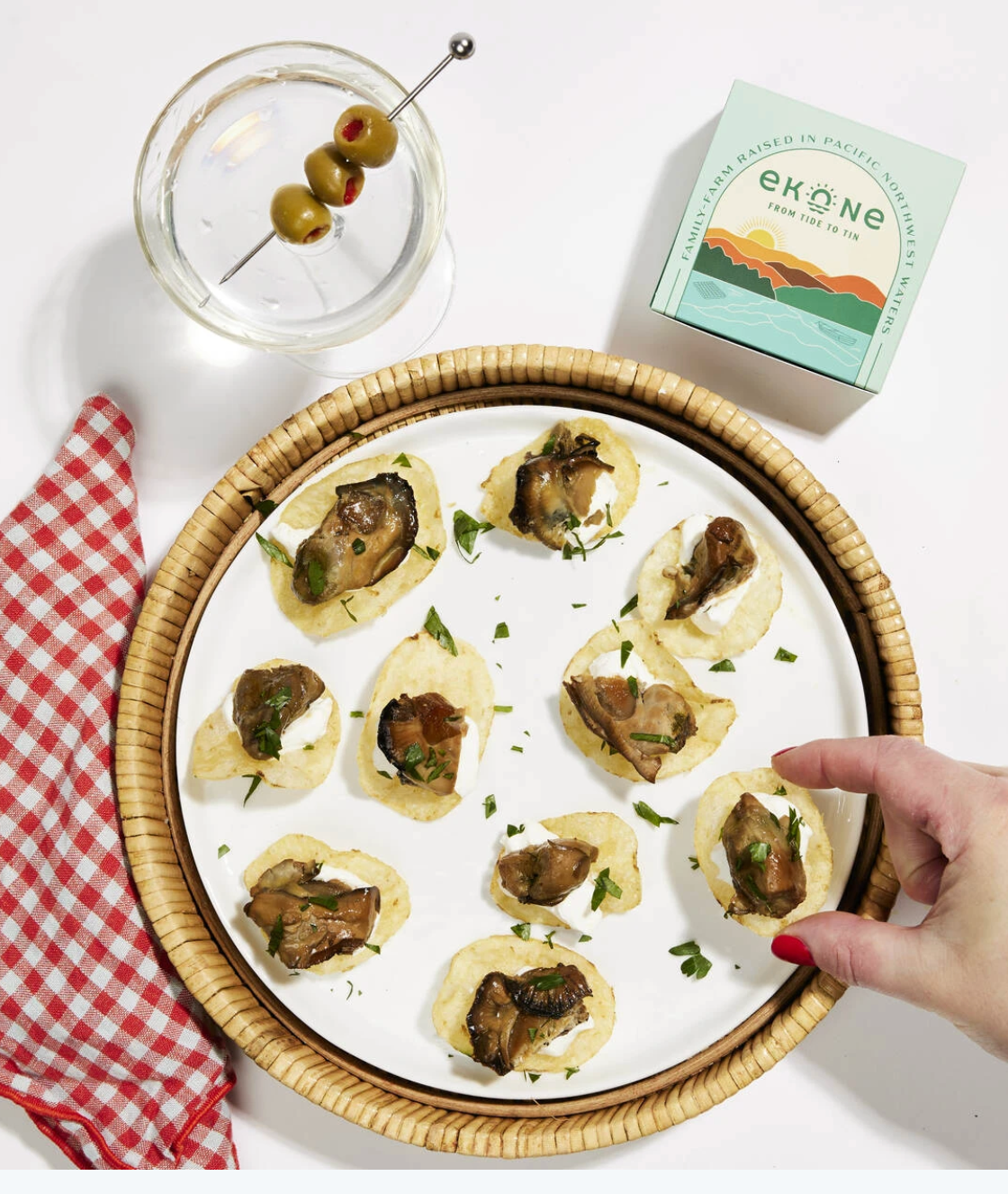 A styled food presentation on a white surface showing several small appetizers on a round white plate with a woven rim, alongside a glass of olives on a toothpick, a red-and-white checkered napkin, and a mint box labeled eköne. A hand is reaching in from the right side.