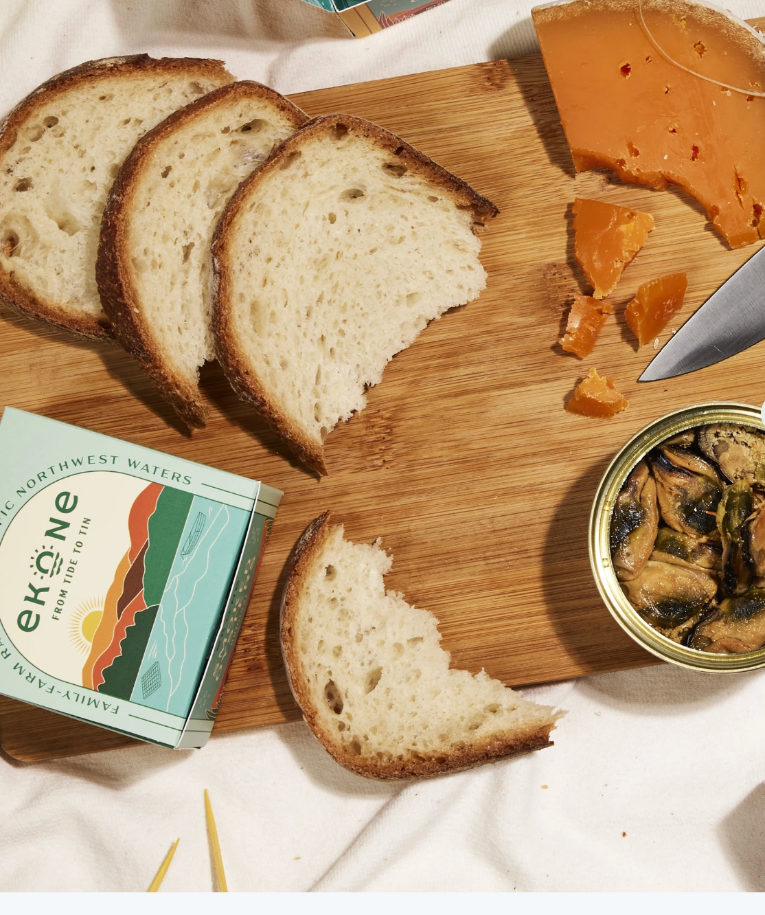A styled food scene on a wooden board with sliced rustic bread, a wedge of orange cheese, a knife, a tin of stuffed mushrooms, and a box labeled ekone. 