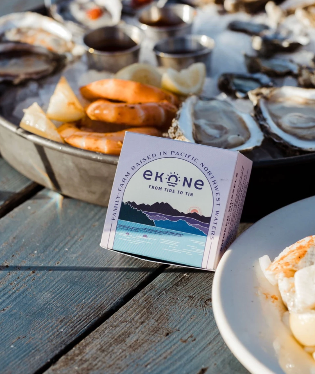 A small Ekone brand tin of Pacific Northwest smoked meats sits on a rustic wooden table surrounded by seafood, including oysters on ice, shrimp, and a plate of food, in an outdoor dining setting.