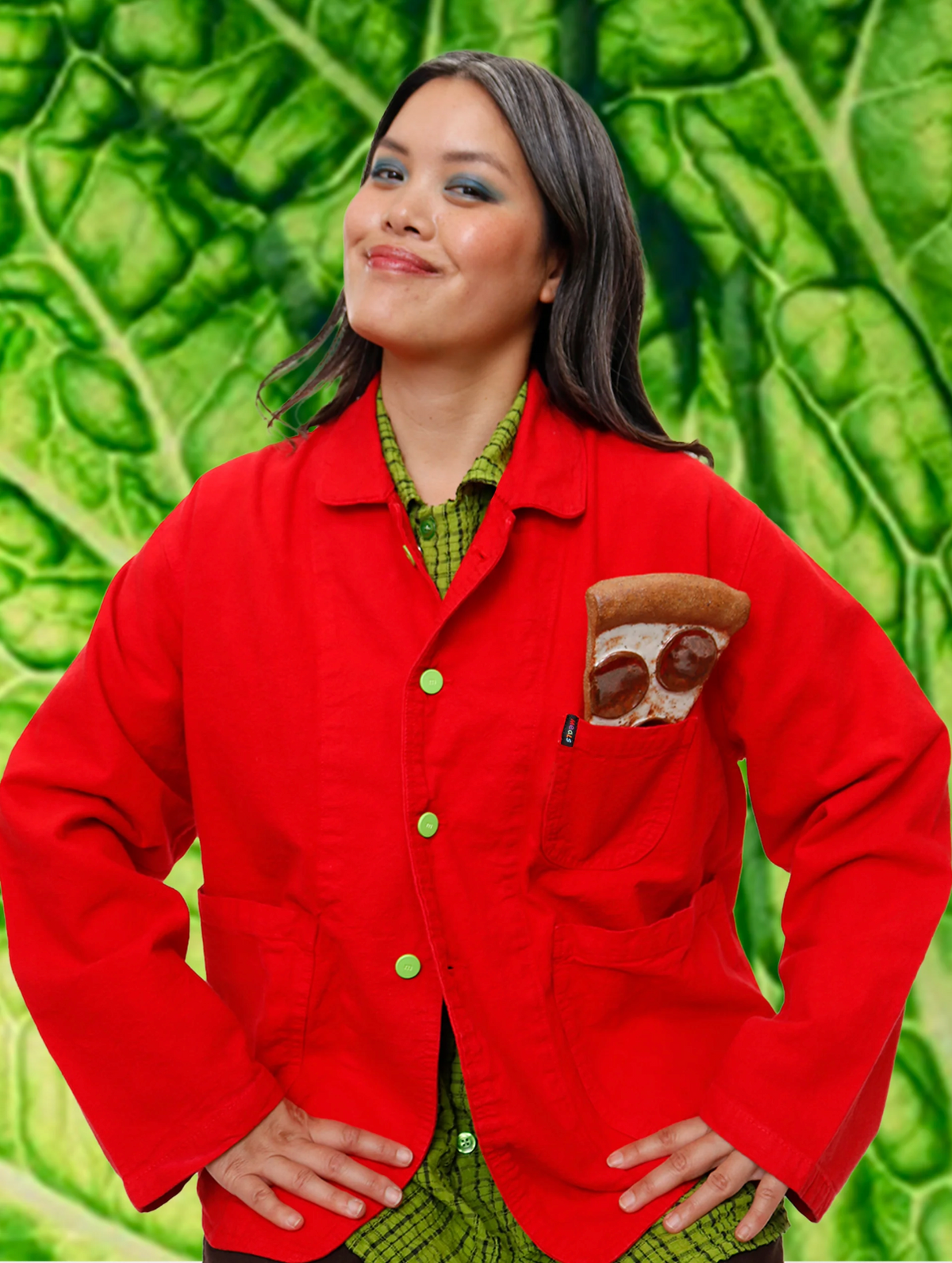 A woman wearing a bright red utility-style jacket with a green plaid shirt underneath, posing with a pepperoni slice in her pocket against a green leafy background.