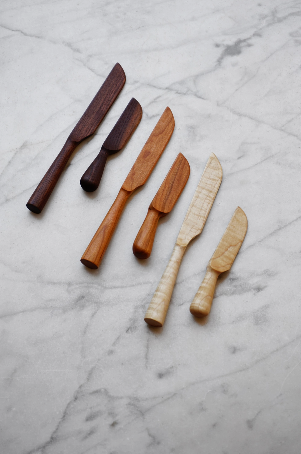 Six handcrafted wooden knife servers in various wood tones, arranged diagonally on a white and gray marble surface.