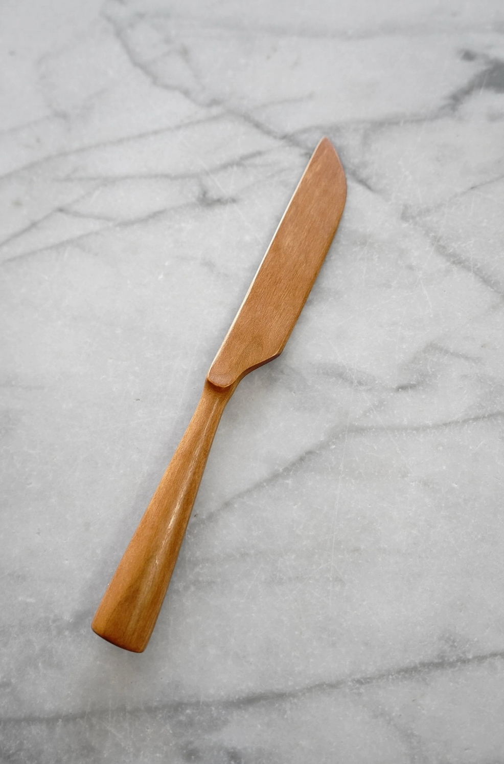 A single wooden knife with a smooth, polished finish and a rounded handle, resting diagonally on a light gray marble surface.