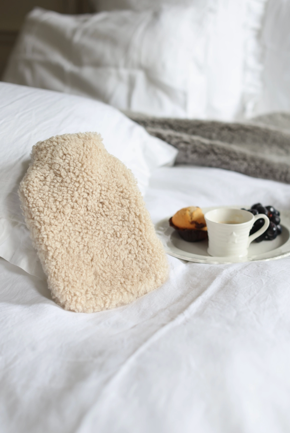 A beige sherpa hot water bottle resting on a white bed next to a small white cup and plate with fruit, in a softly lit bedroom setting.