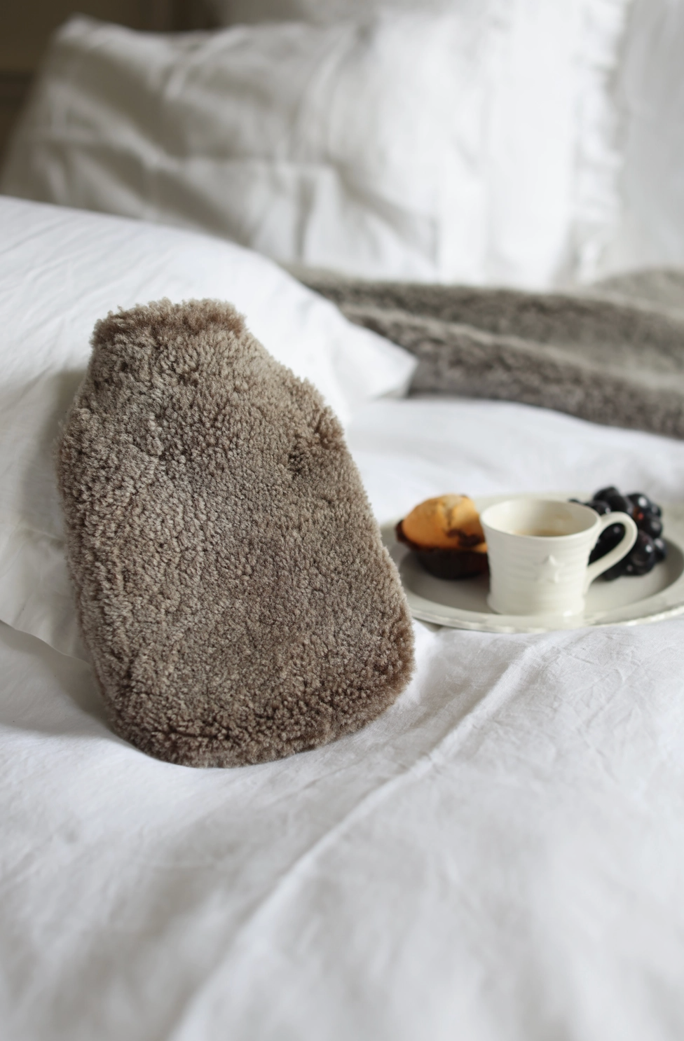 A brown textured hot water bottle with fluffy faux fur, resting on a white bed next to a plate with a cup and fruit.