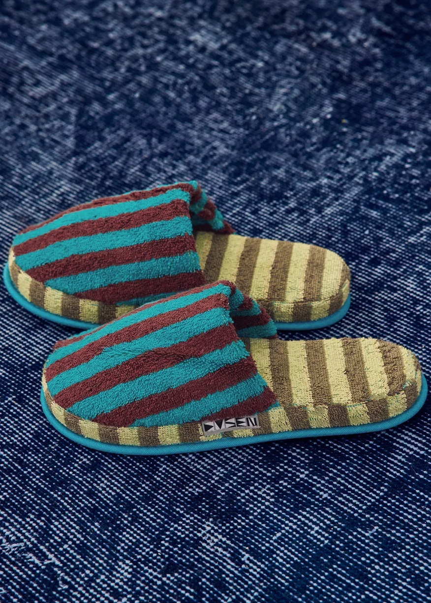 A pair of plush slip-on slippers with horizontal stripes in teal, burgundy, and tan, displayed on a textured dark blue denim-like fabric background.