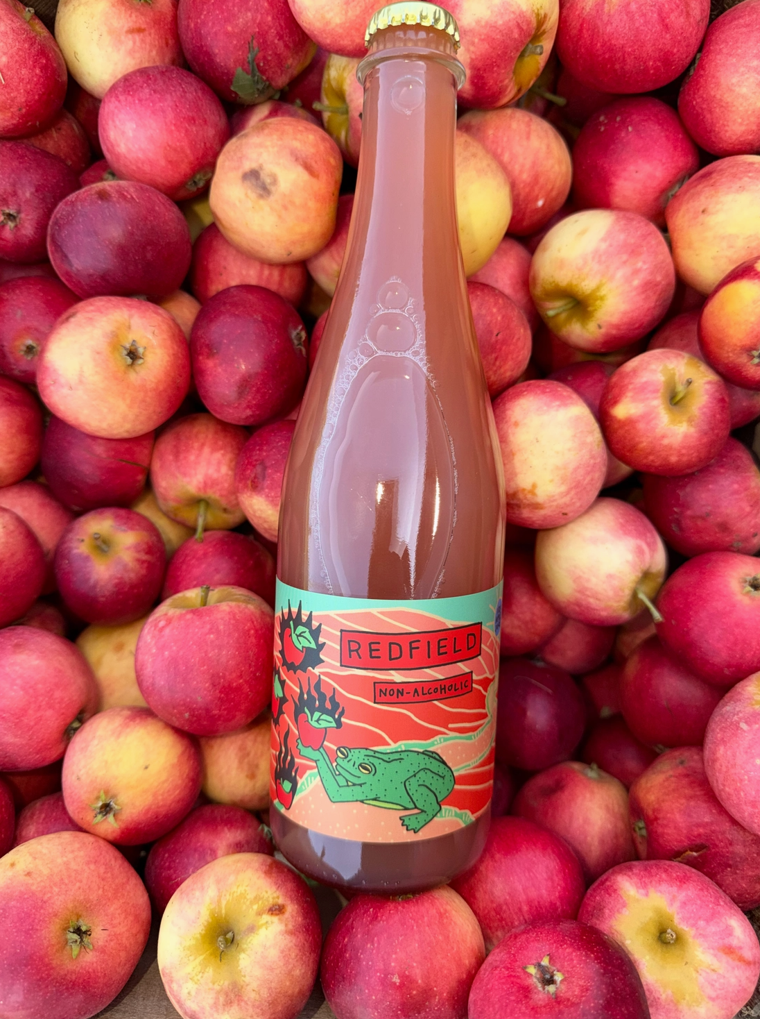A bottle of Redfield filled with pinkish-red liquid, set against a background of fresh red and yellow apples. The bottle features a colorful label with a green frog illustration and the brand name 'Redfield'.
