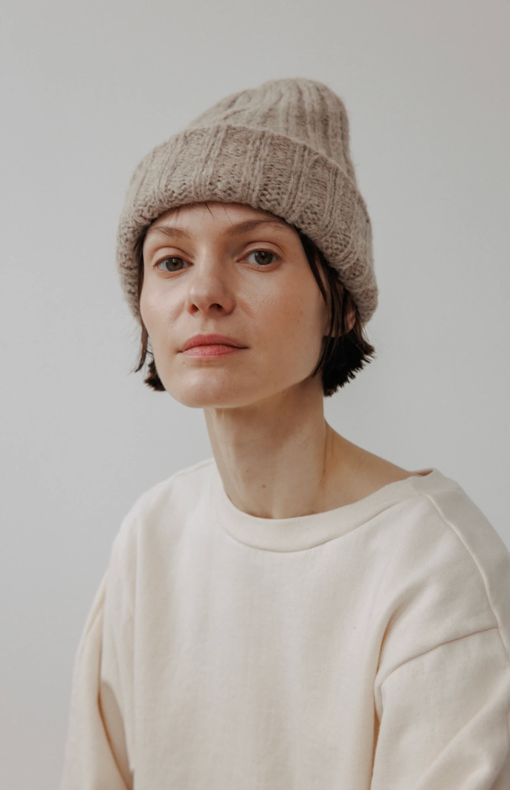 A person wearing a beige chunky knit beanie hat and a cream-colored sweatshirt, photographed against a plain light gray background.