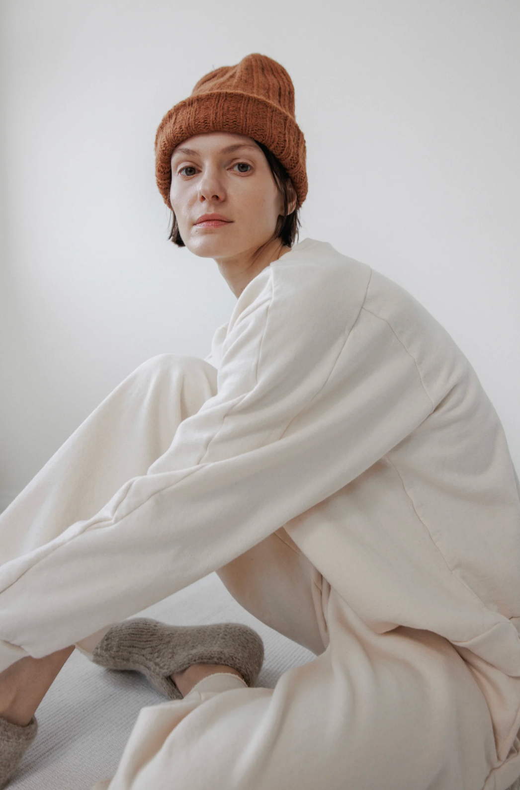 A person wearing a rust-colored knit beanie, a cream-colored lounge set, and fuzzy gray slippers, sitting against a plain light gray background.