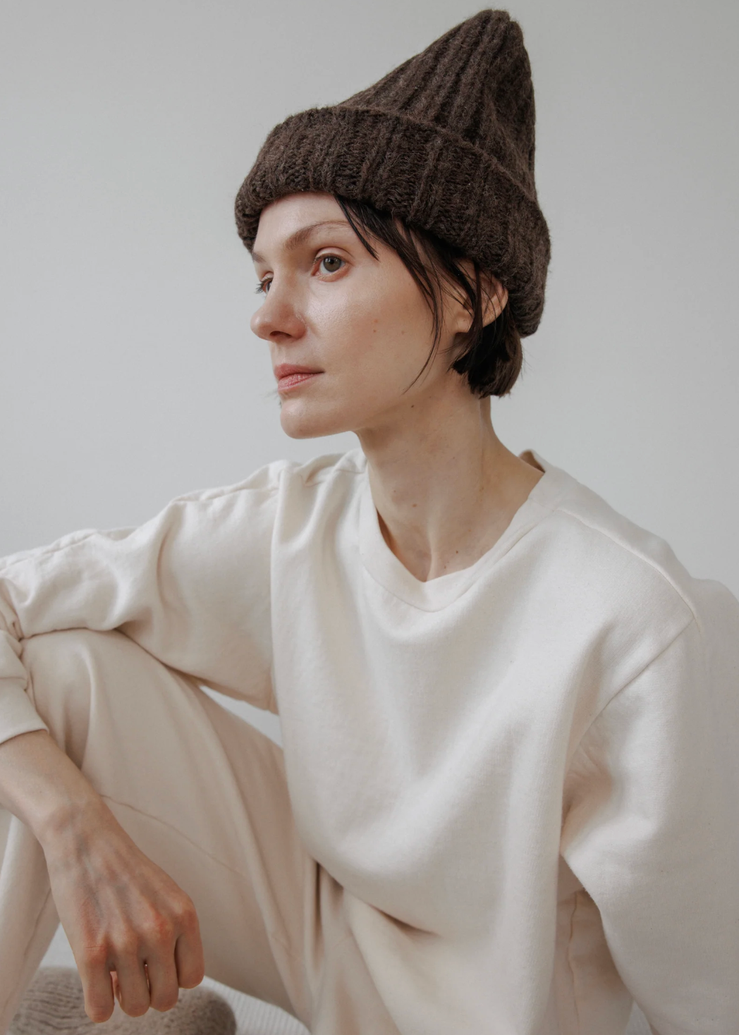 A person sitting on a white textured floor against a white wall, wearing a cream-colored lounge set consisting of a sweatshirt and pants, a brown knit beanie, and a beige knit slipper.