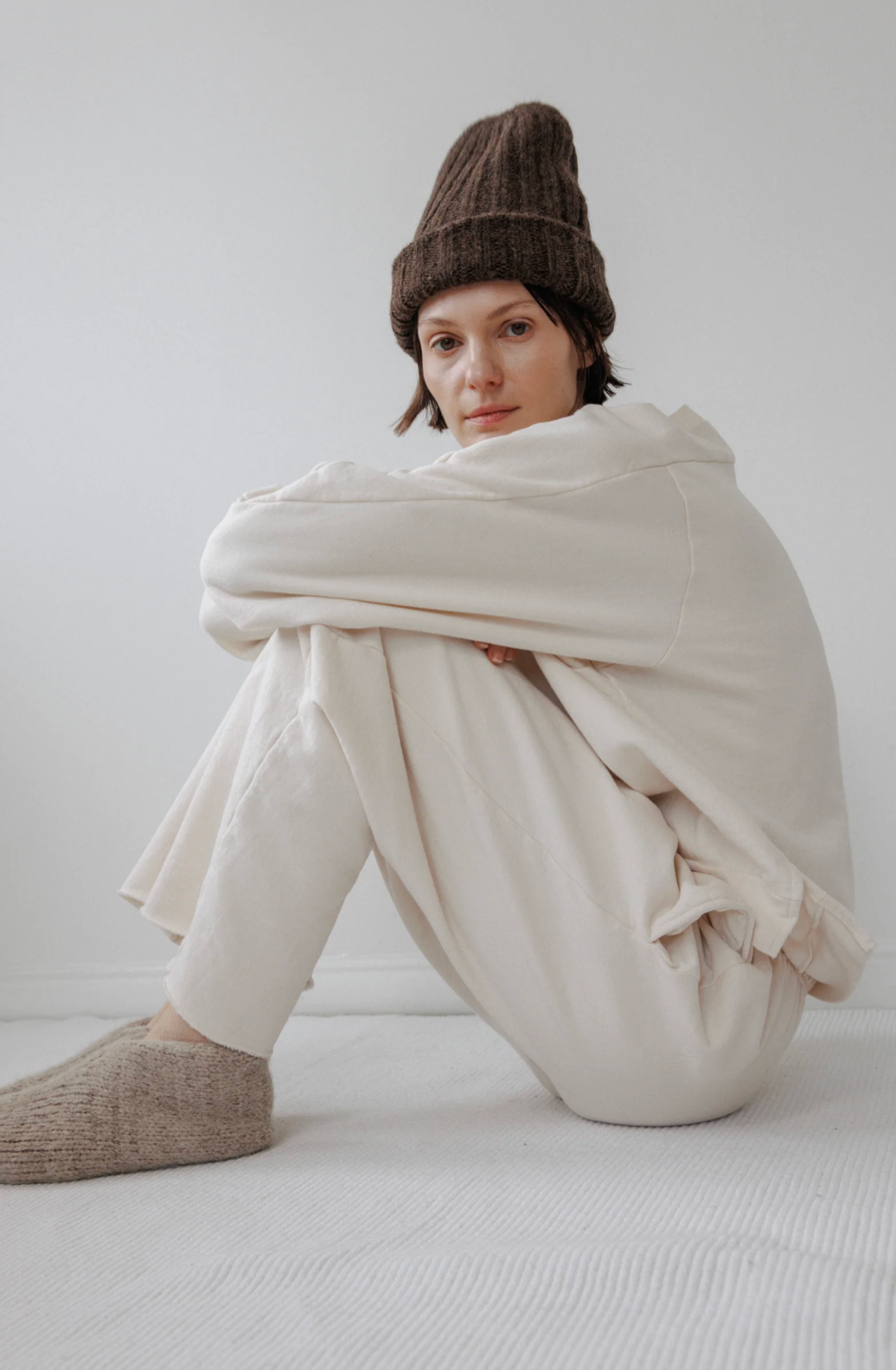A person sitting on a white textured floor against a white wall, wearing a cream-colored lounge set consisting of a sweatshirt and pants, a brown knit beanie, and a beige knit slipper.