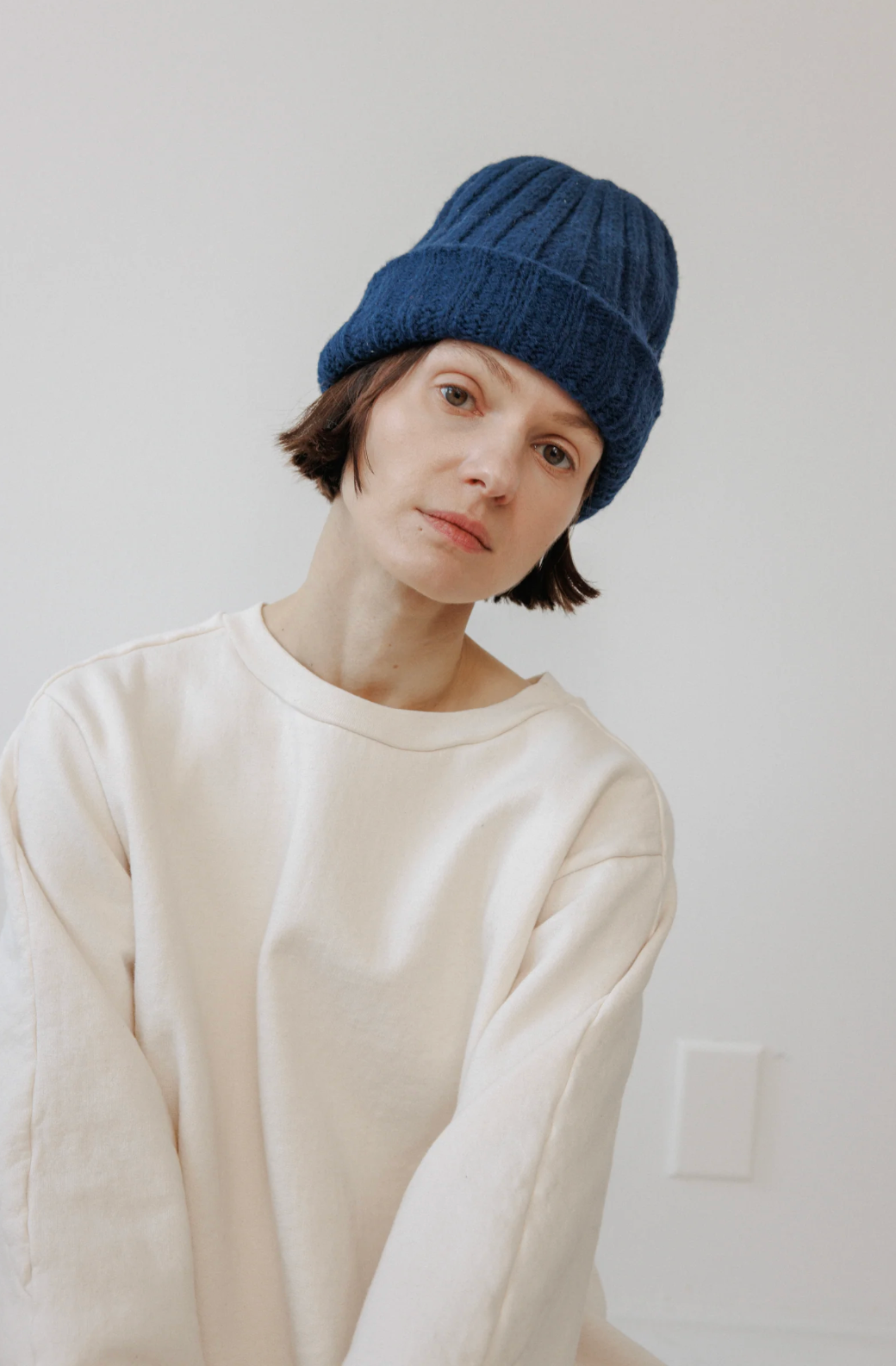 A person wearing a navy blue ribbed knit beanie and an oversized cream-colored long-sleeve top, photographed against a plain off-white wall.