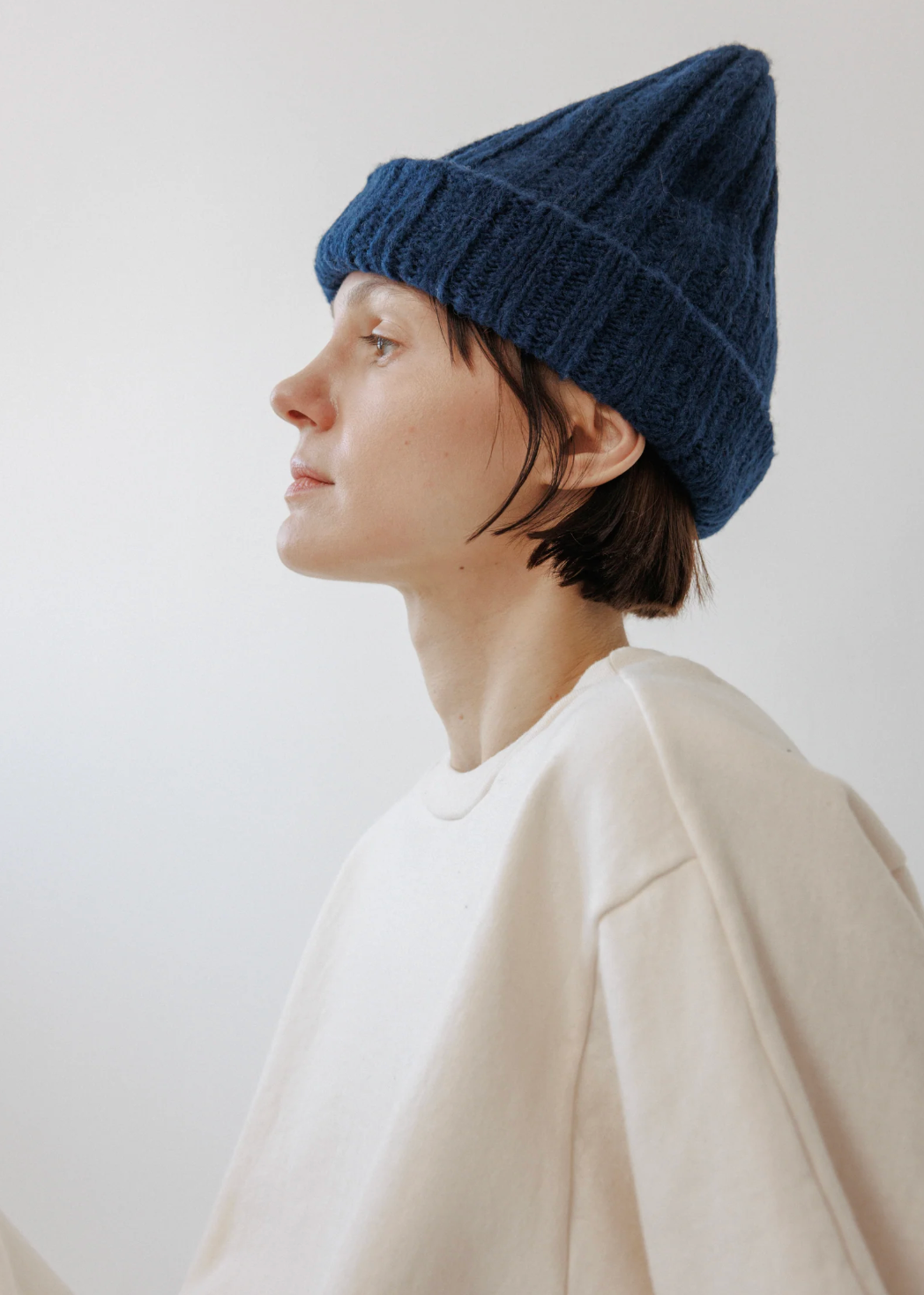 A person in profile wearing a dark navy blue ribbed knit beanie and a cream-colored sweatshirt, photographed against a plain light gray background.