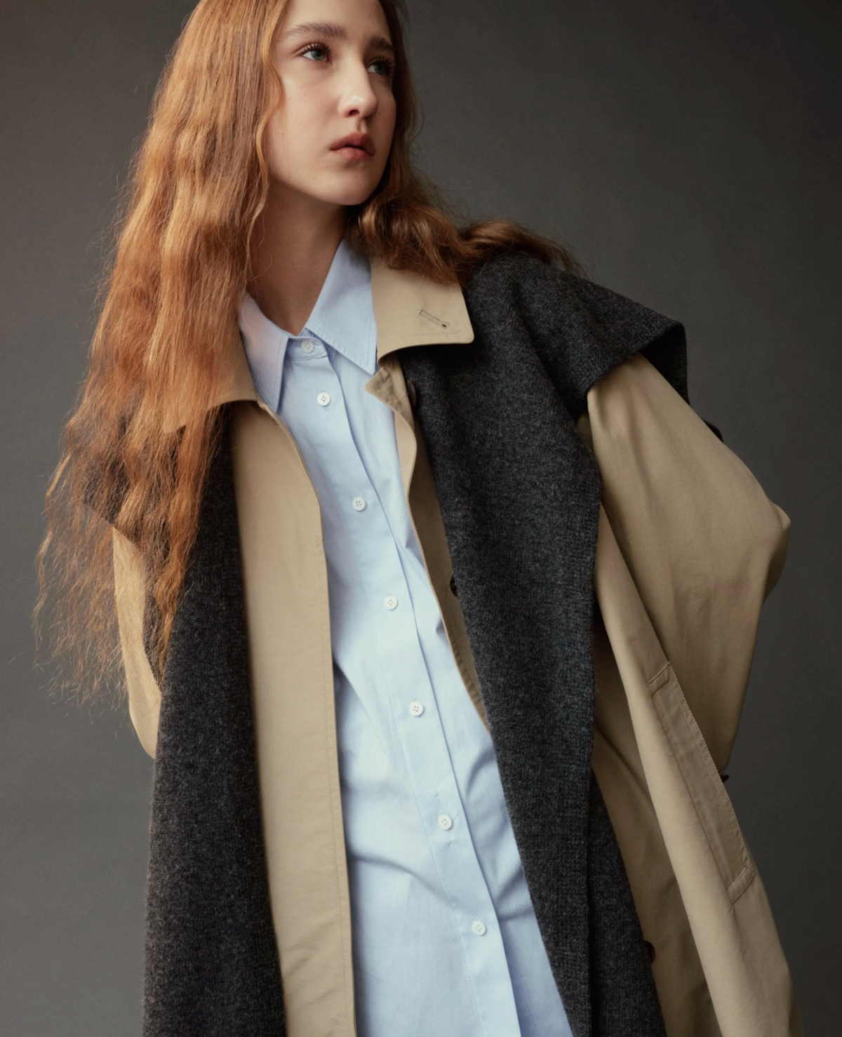 A model with long wavy red hair is shown from the chest up, wearing a light blue button-down shirt layered under a structured beige and dark gray coat against a dark gray studio background.