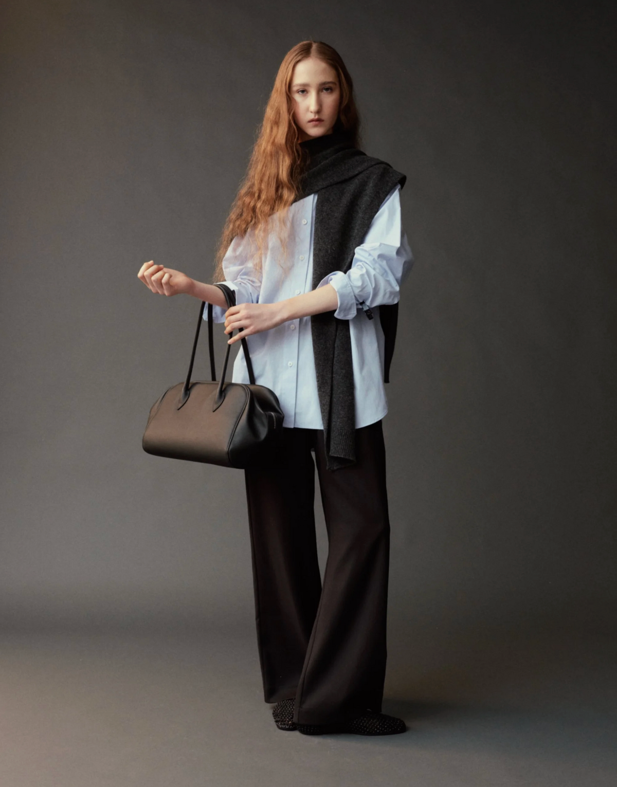 A model stands against a dark gray studio background wearing a white shirt, black wide-leg trousers, a black knit shawl, and holding a black leather handbag.